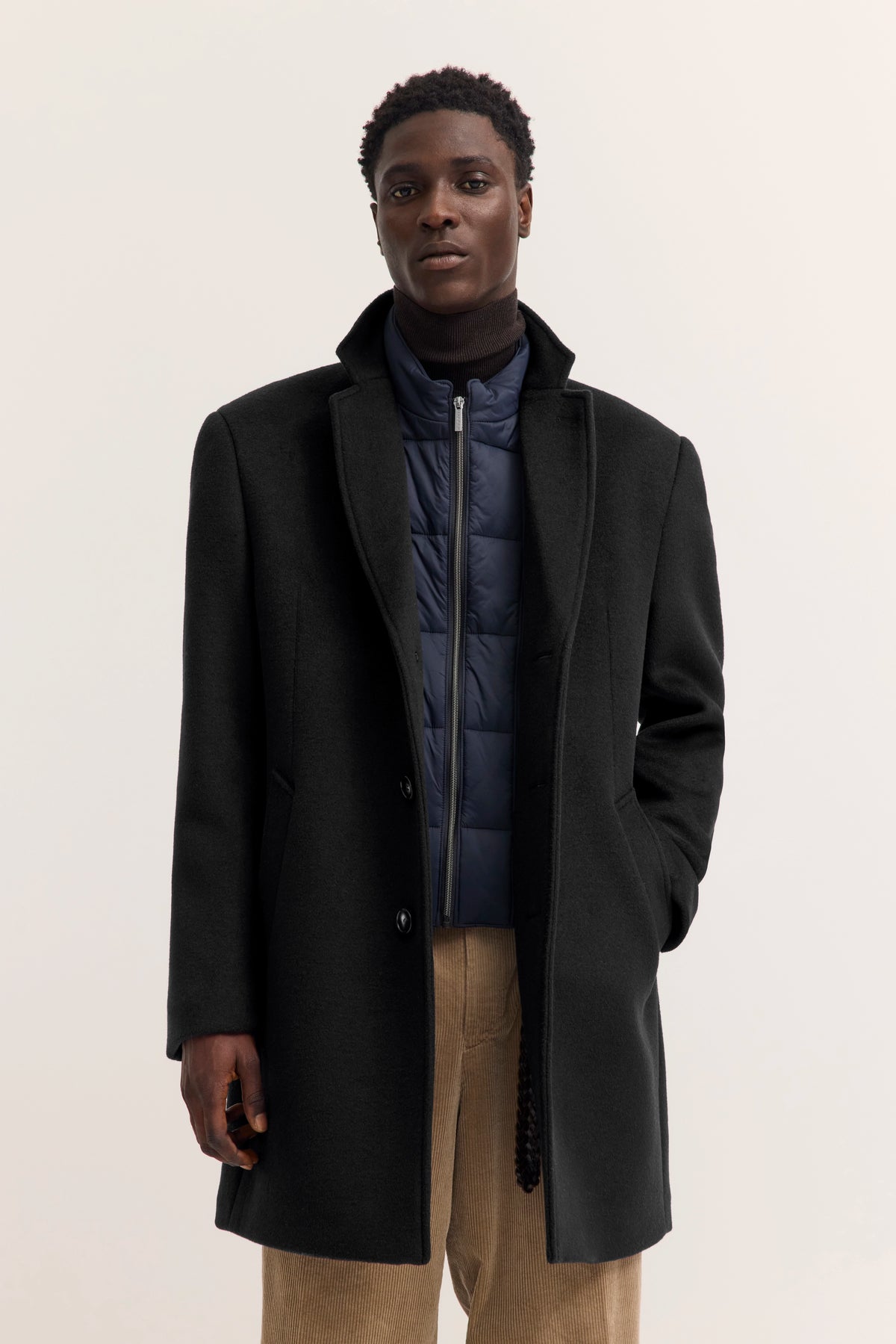 Modelbild: Schwarzer Mantel, blauer Steppjacke, beige Hose. Entspanntes, selbstbewusstes Posing.