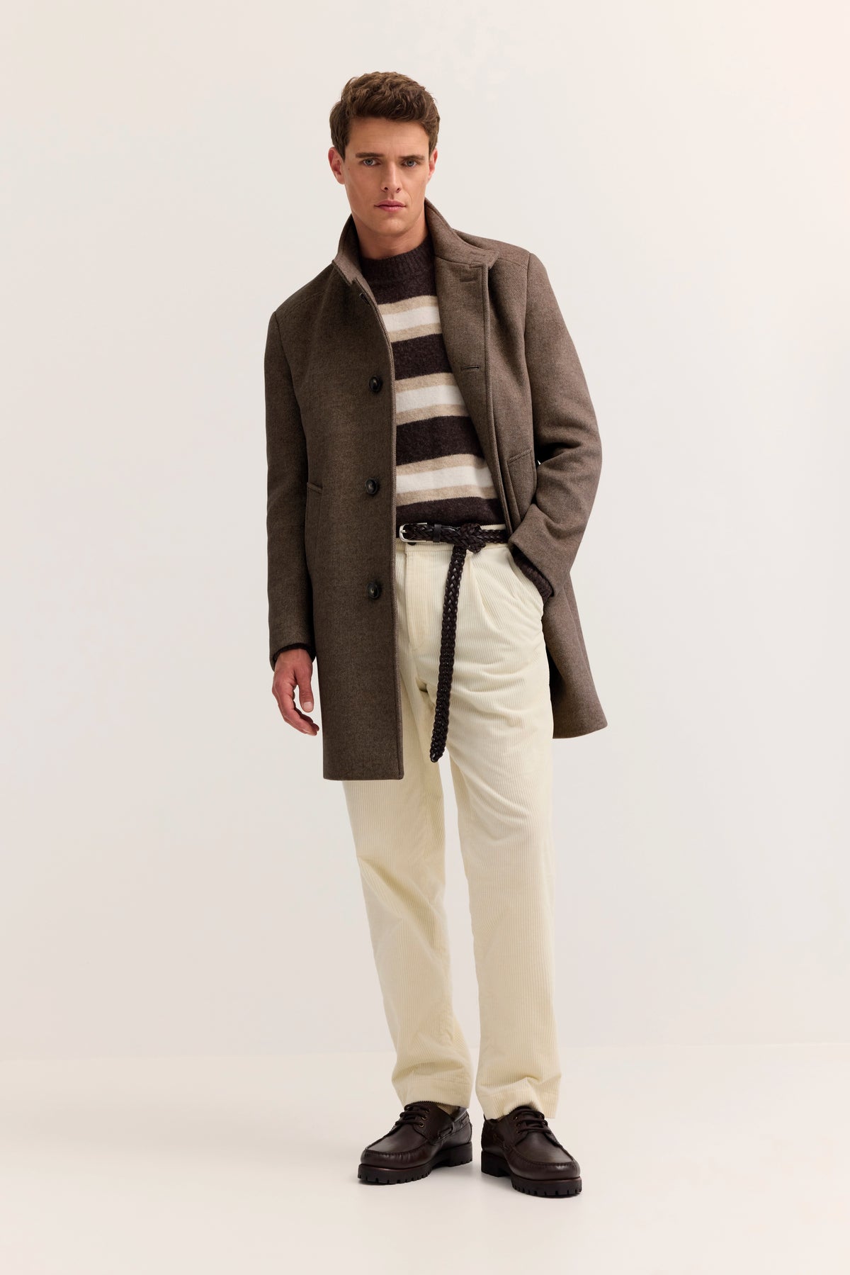 Brauner Mantel, gestreifter Pullover, helle Hosen. Model steht locker mit Hand in der Tasche.
