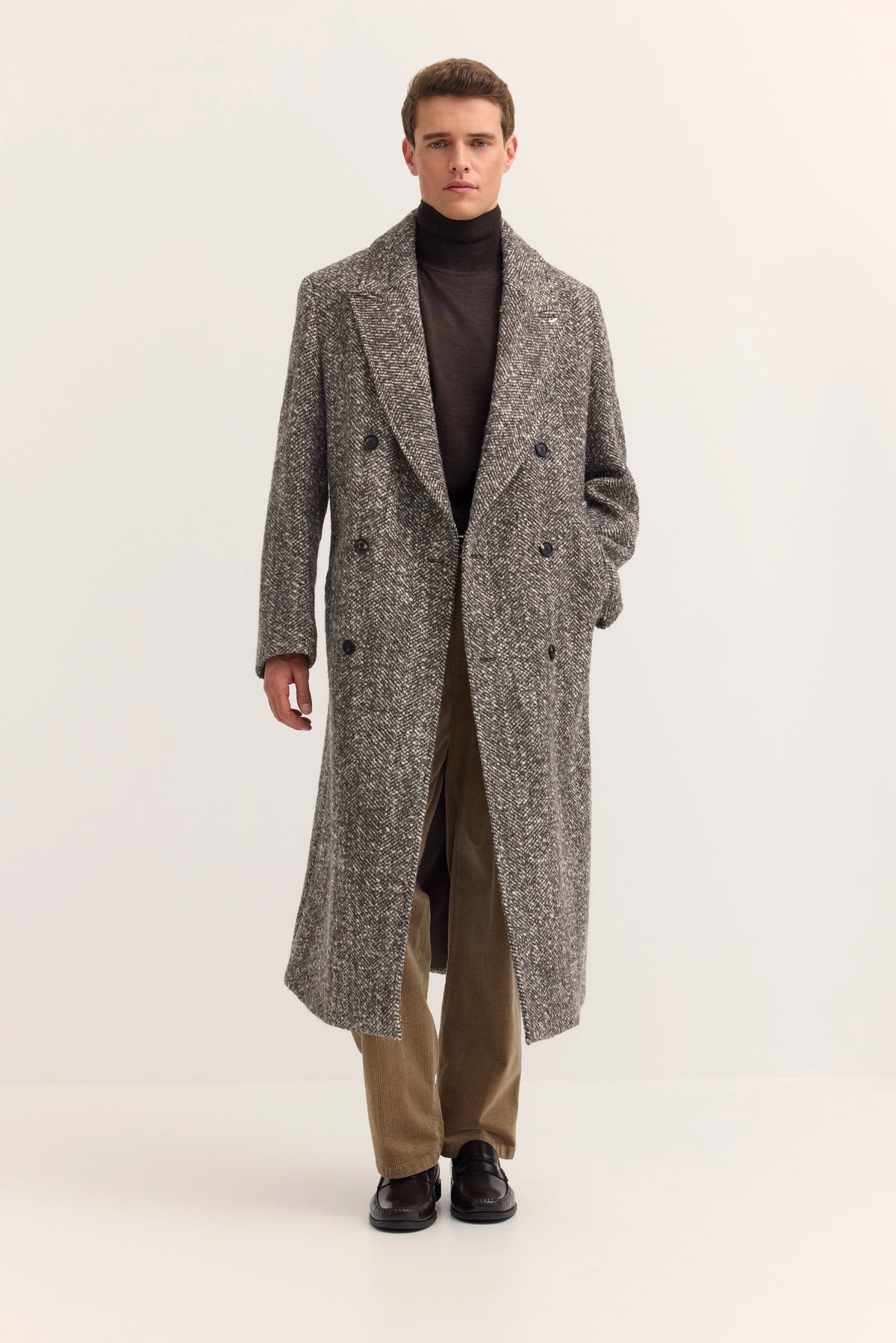 Model in grauem Mantel, braunem Rollkragenpullover und beigen Hosen. Lässig elegant, stehend.
