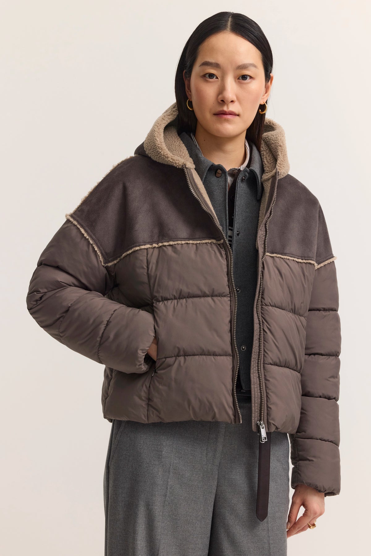 Damen Winterjacke, braun, mit Kapuze und Schafsfellfutter, über grauem Hemd, Modelbild, entspannte Pose.