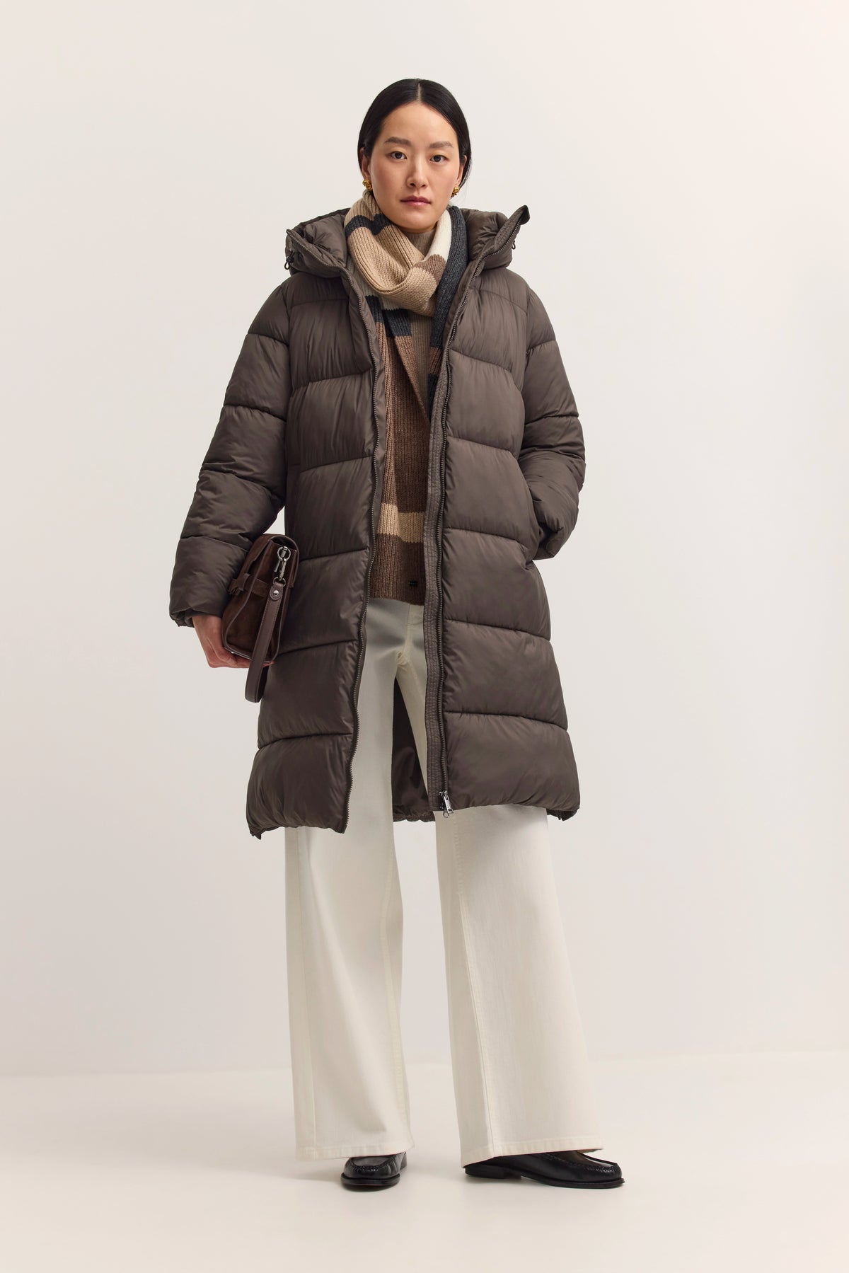 Model in braunem bugatti Daunenmantel, beigem Pullover und weißer Hose, legeres Stehposing.