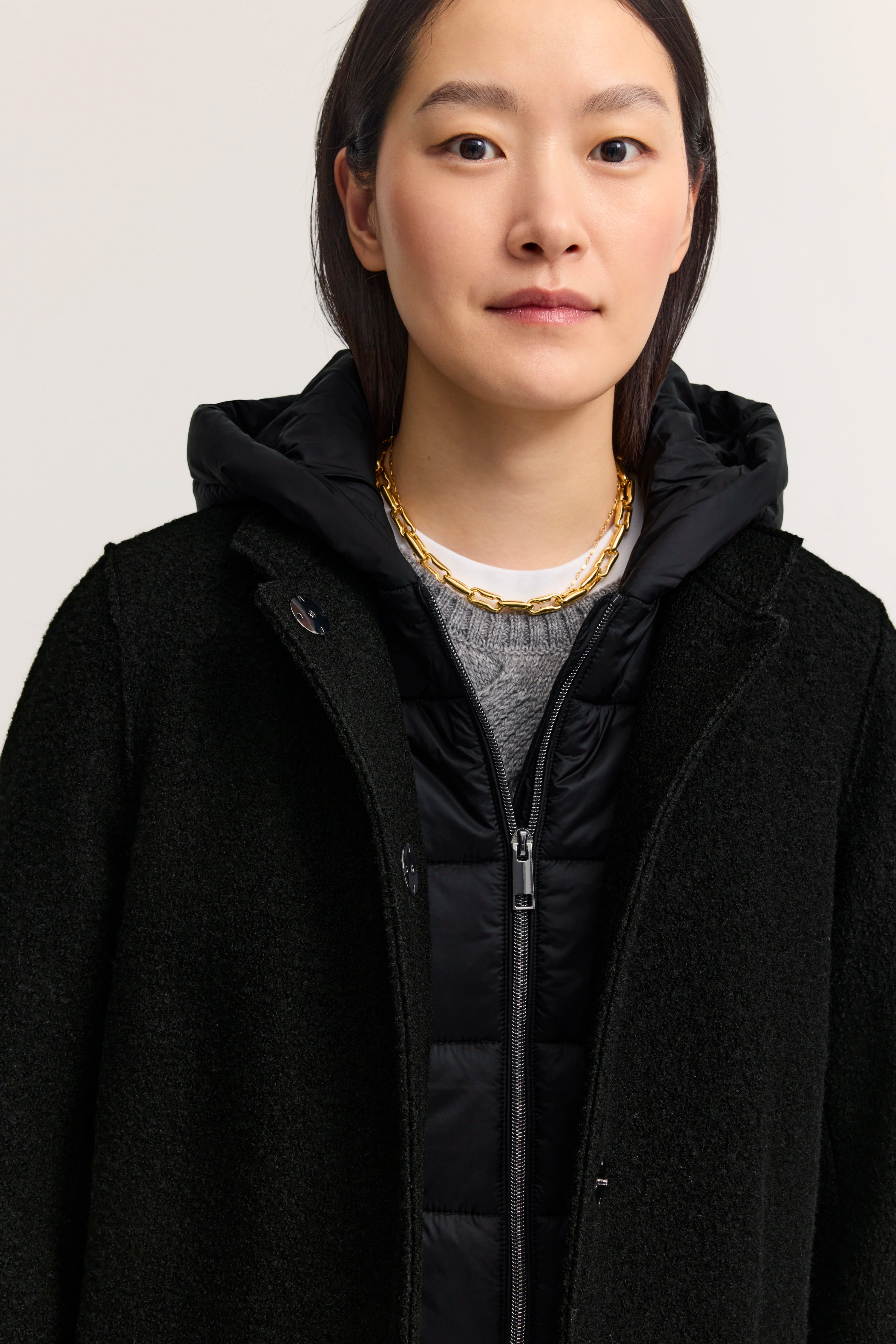 Schwarzer Mantel über schwarzer Steppjacke, grauer Pullover, Modelbild, neutraler Gesichtsausdruck.