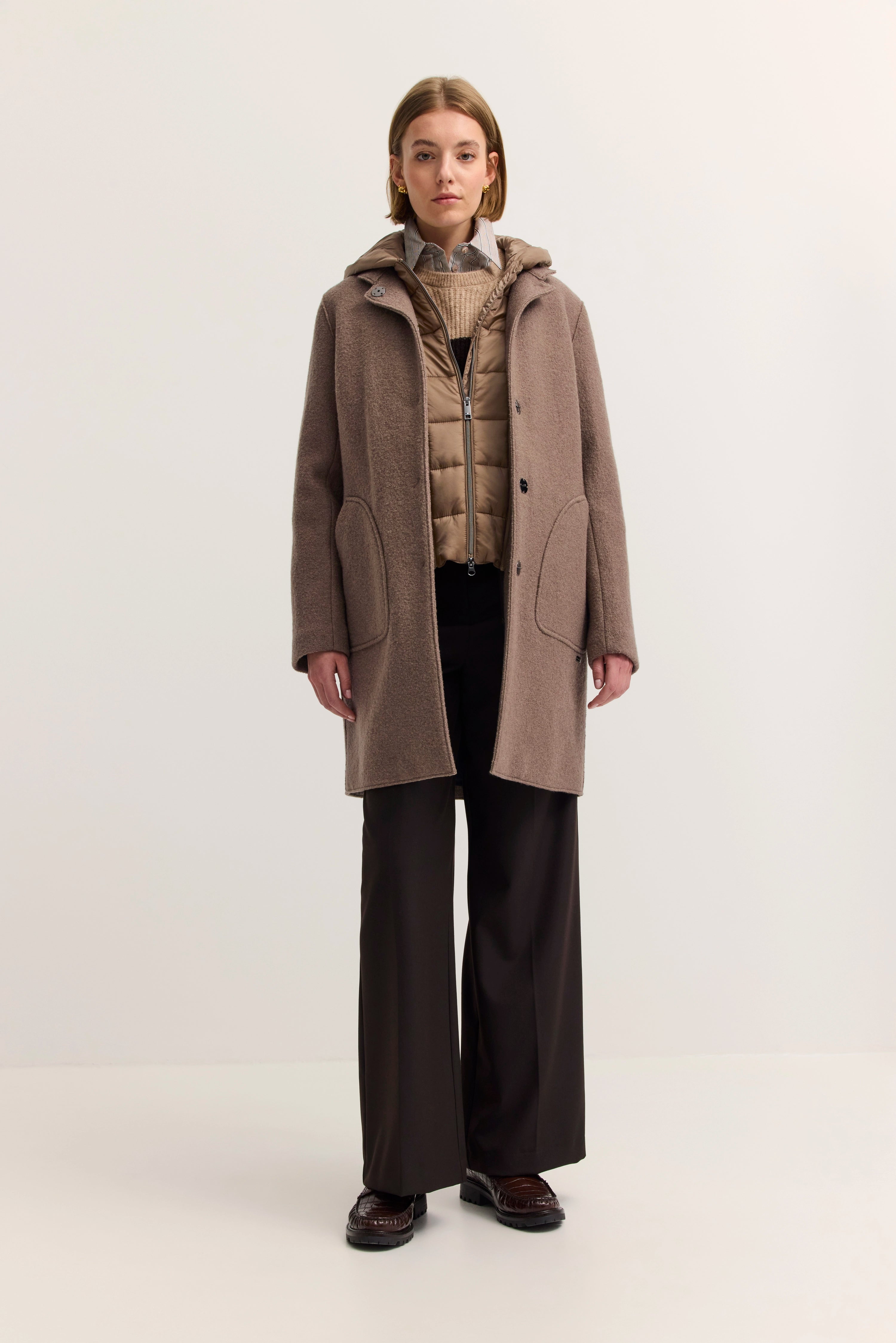 Model in taupefarbenem bugatti-Mantel und Daunenweste, darunter Strickpullover und gestreiftes Hemd.