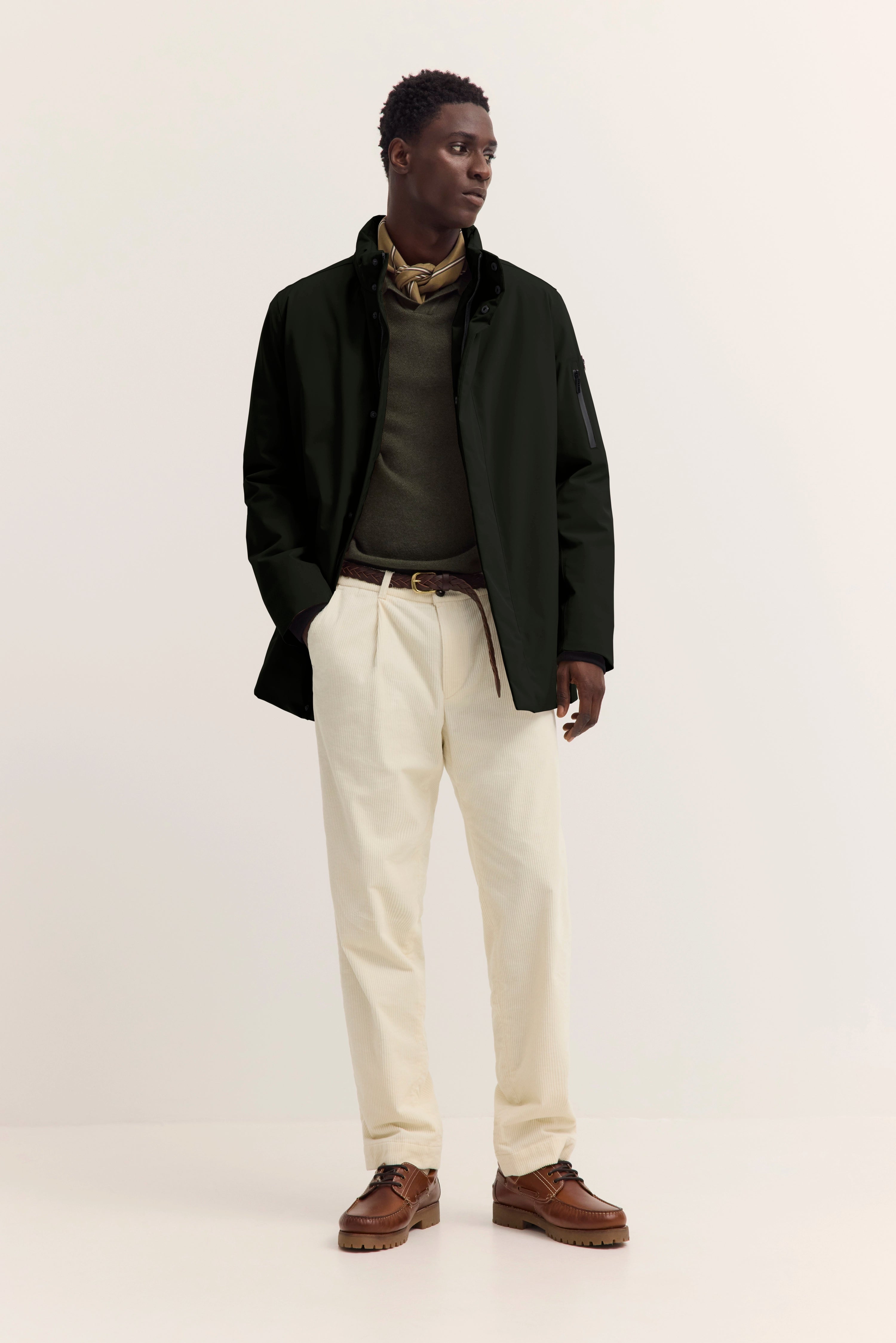 Model in schwarzer bugatti-Jacke, olivgrüner Pullover, beiges Halstuch. Legeres Posing.