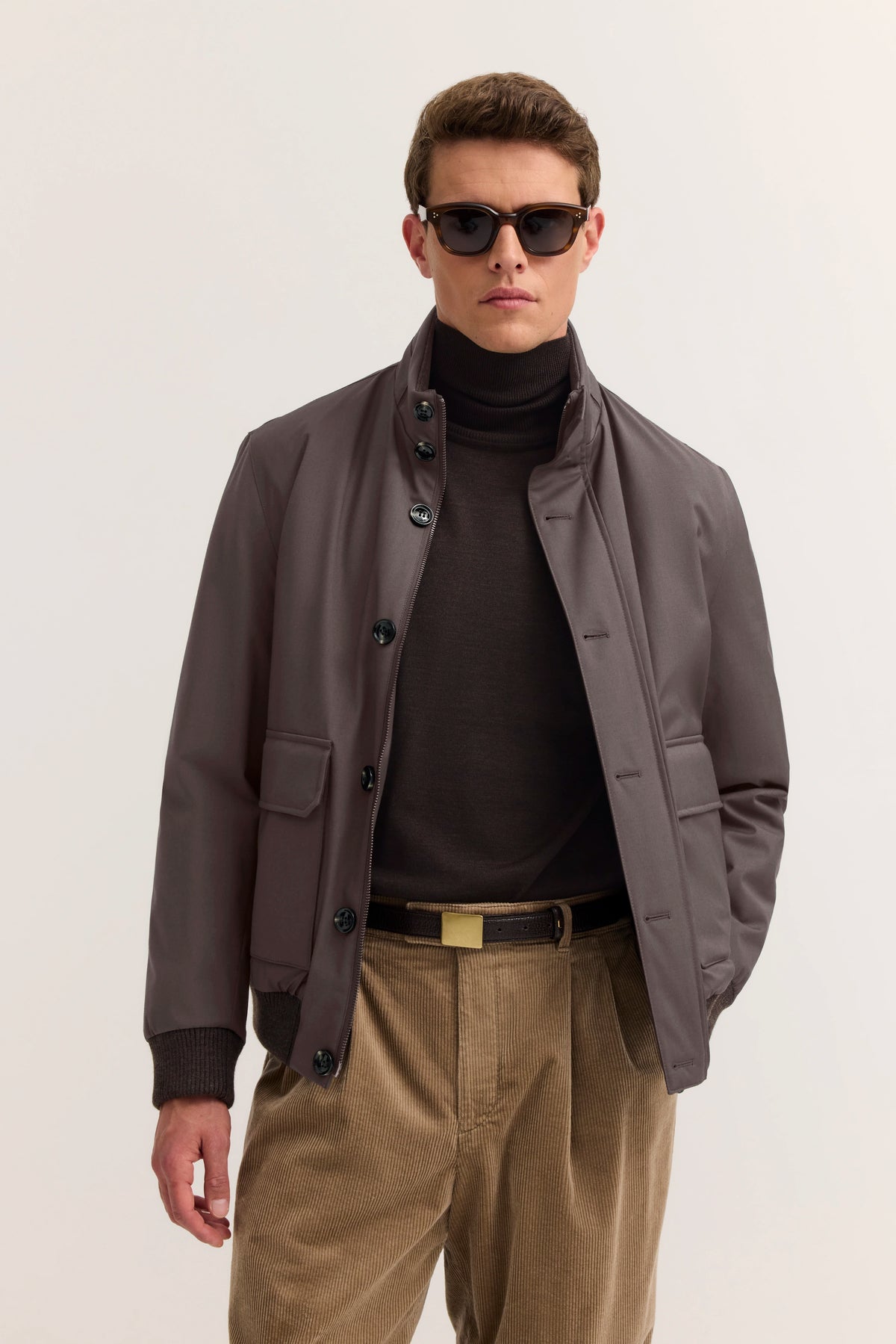 Model in brauner Jacke und Rollkragenpullover, beigefarbene Cordhose, Hände in den Taschen, entspannte Pose.