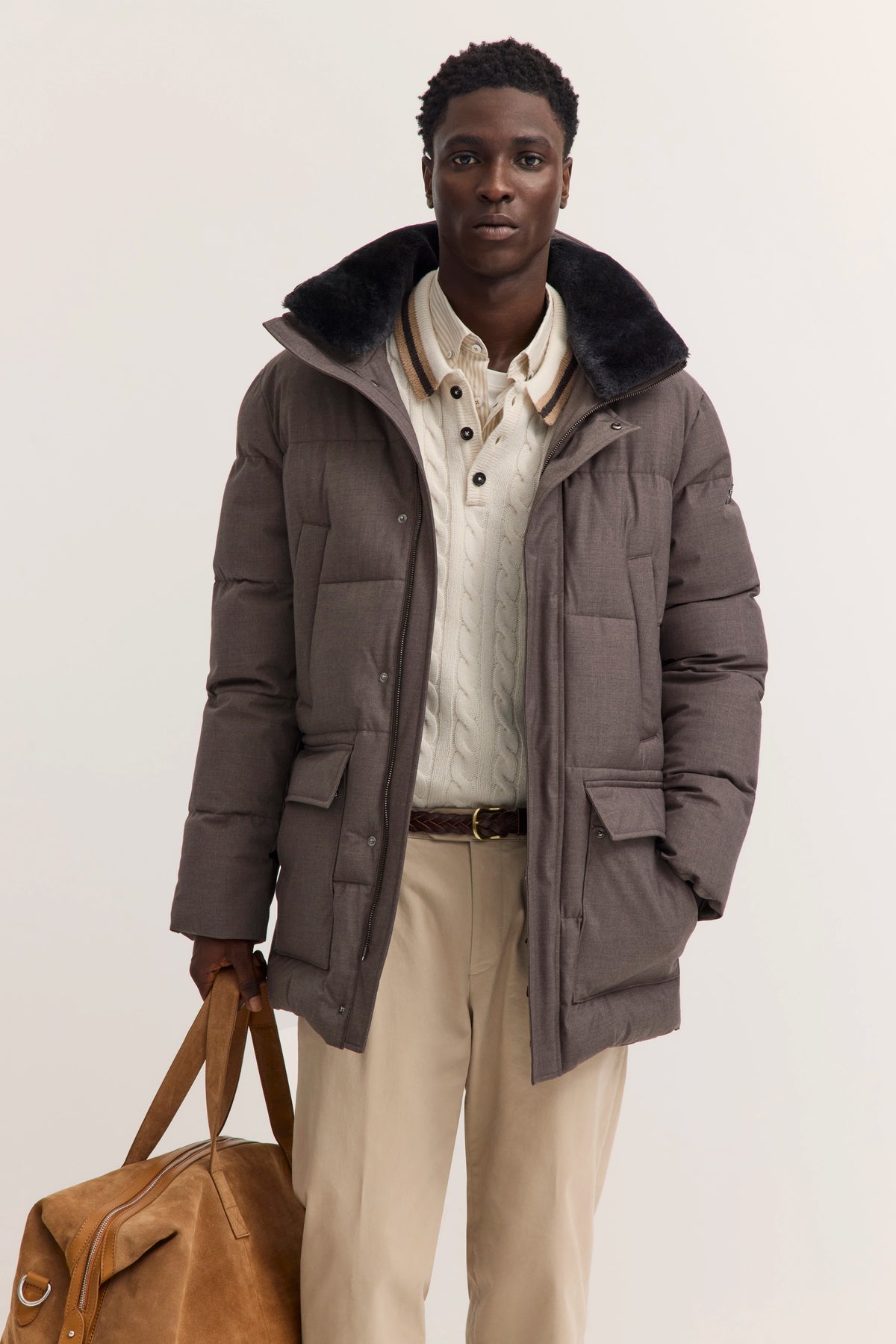 Model trägt graue bugatti Daunenjacke, cremefarbenen Pulli und beige Hose. Entspanntes Posing mit Tasche.