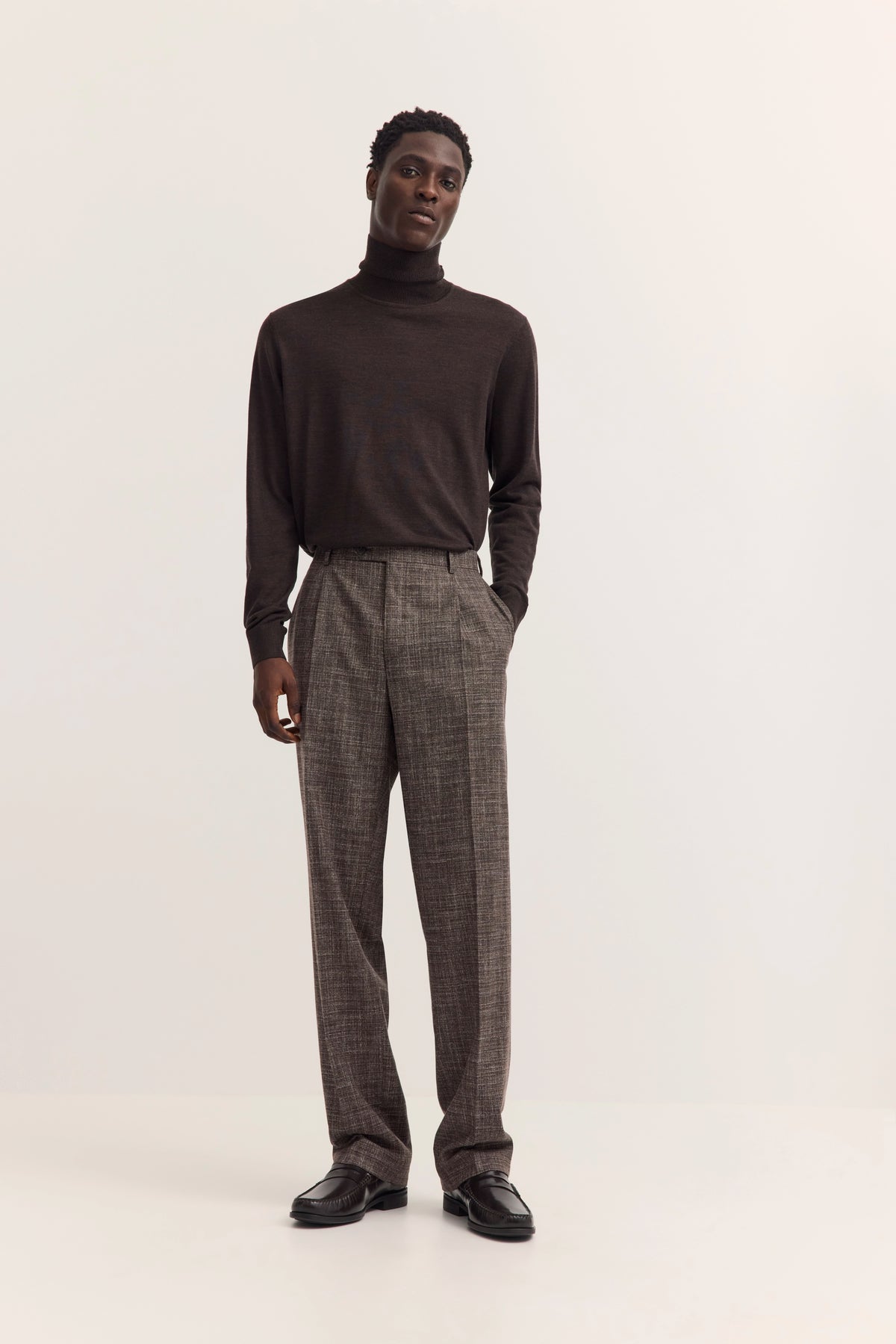 Model in braunem Rollkragenpullover und grauer Hose, lässig stehend.