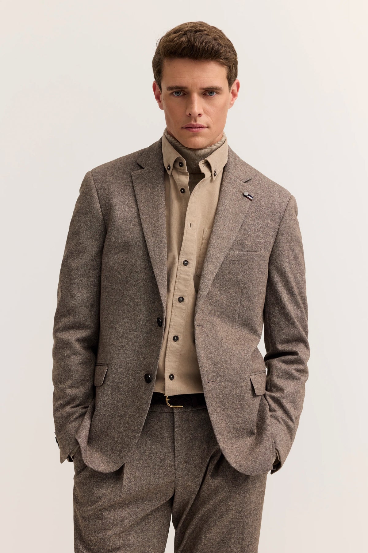Model in braunem Anzug mit beigem Hemd und Rollkragen, Hände in Taschen, bugatti Stil.