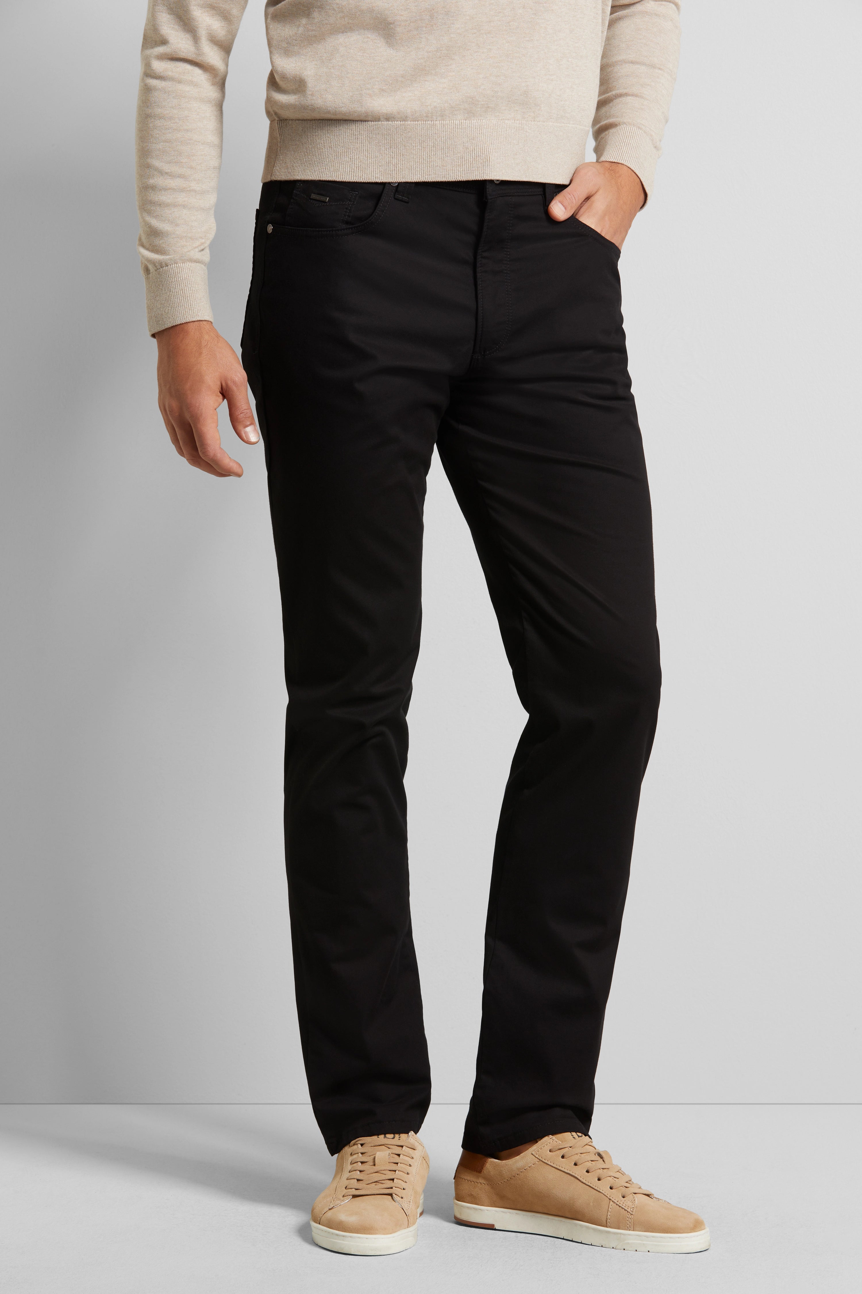 Modelbild in schwarzer Hose und beigem Pullover, lässig stehend, Hände in Hosentaschen.