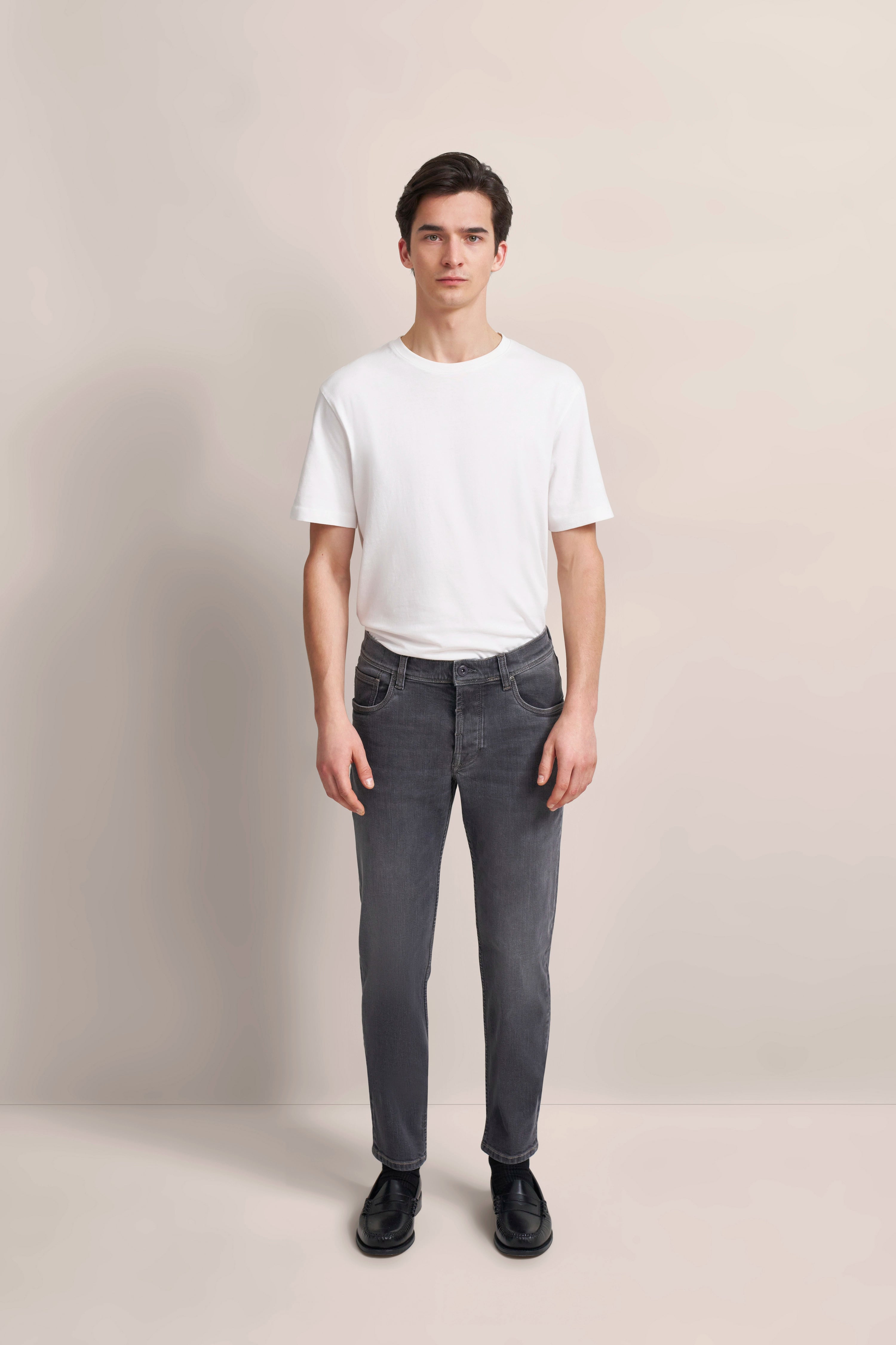Weißes T-Shirt, graue Jeans, schwarze Schuhe. Modelbild, entspannt stehende Pose. bugatti Stil.