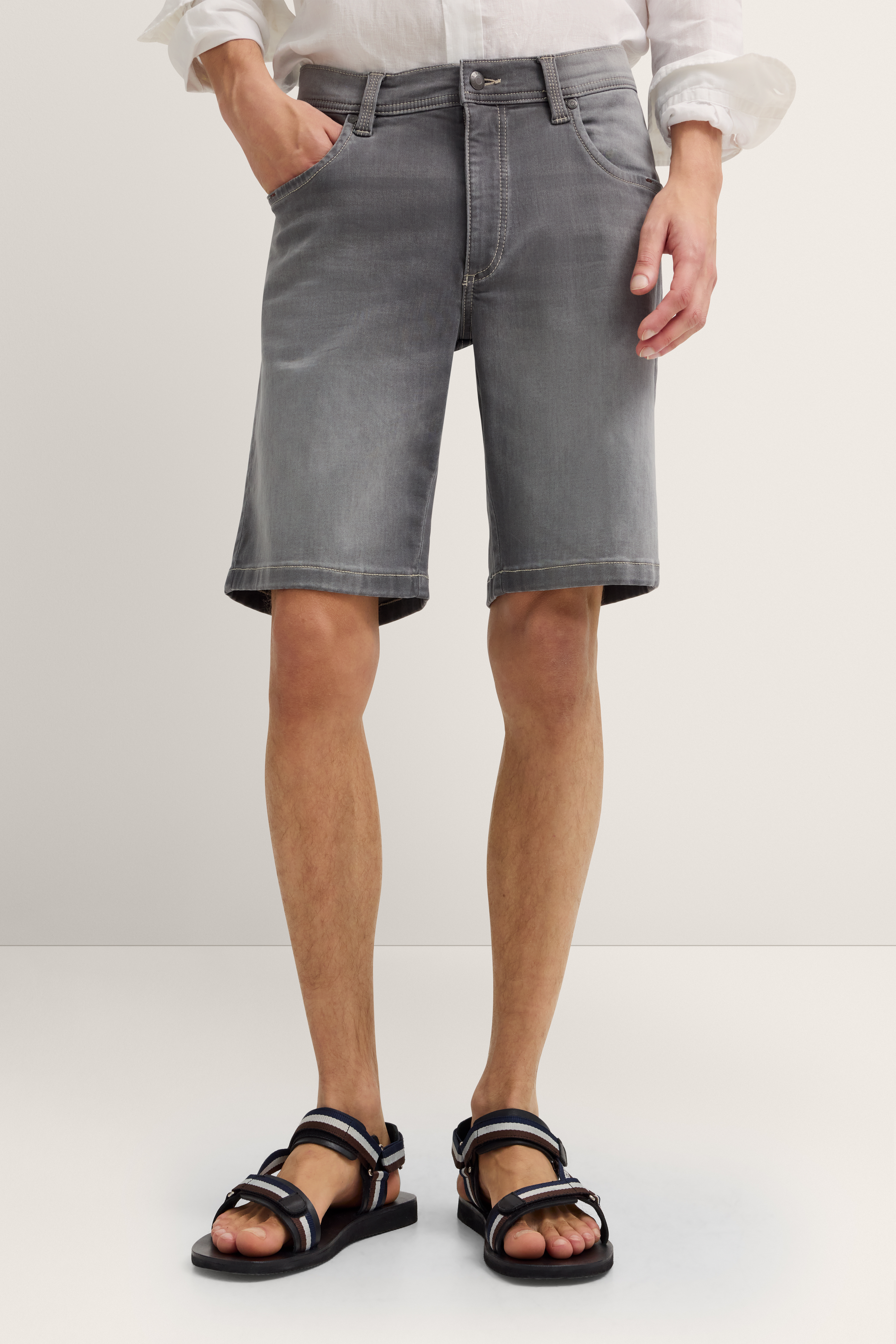 Graue Jeansshorts, weißes Hemd, schwarze Sandalen. Modelbild, Hände in Taschen, entspanntes Posing.