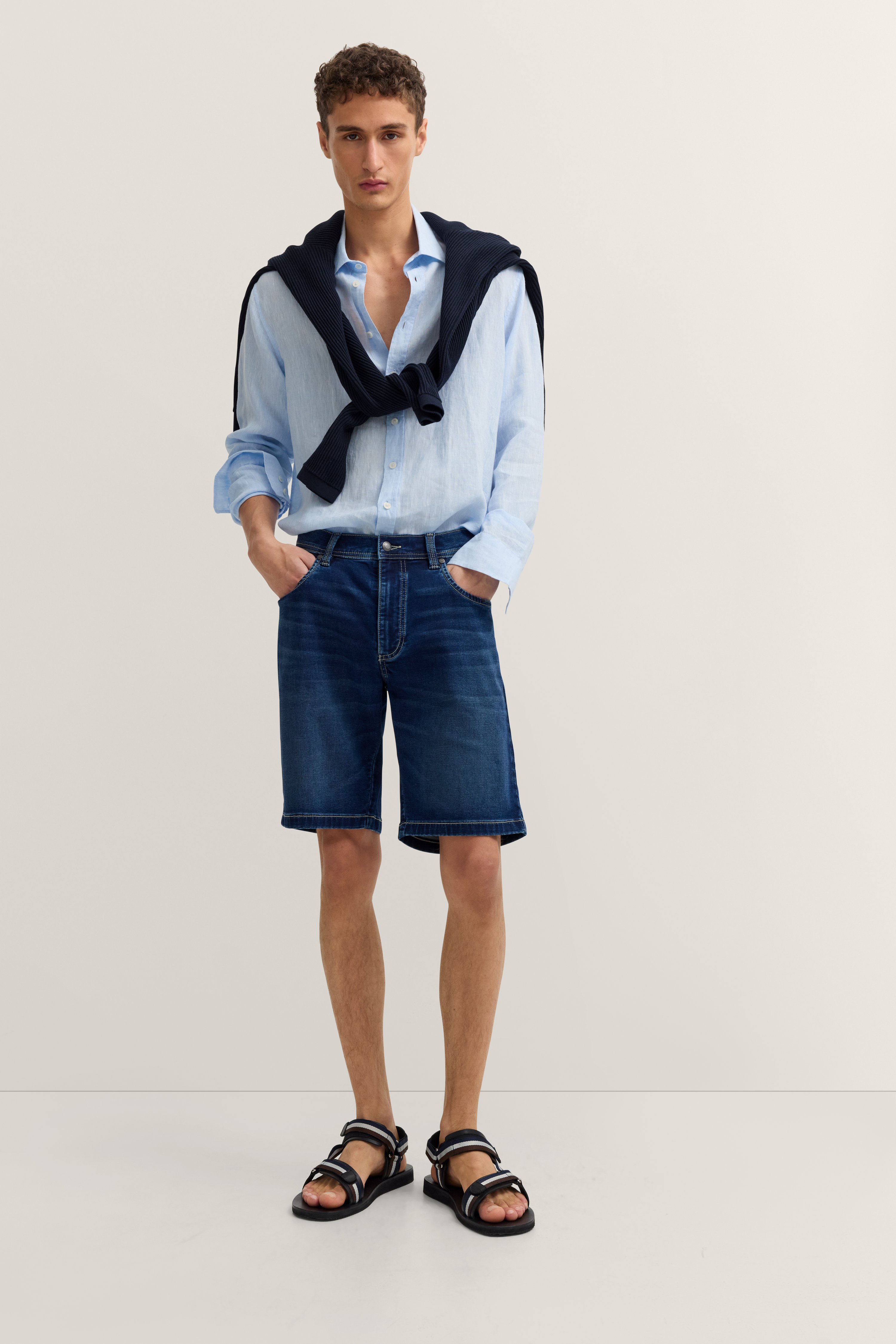 Model in hellblauem Hemd, Jeansshorts, schwarzem Pullover um die Schultern, schwarze Sandalen, entspannte Pose.