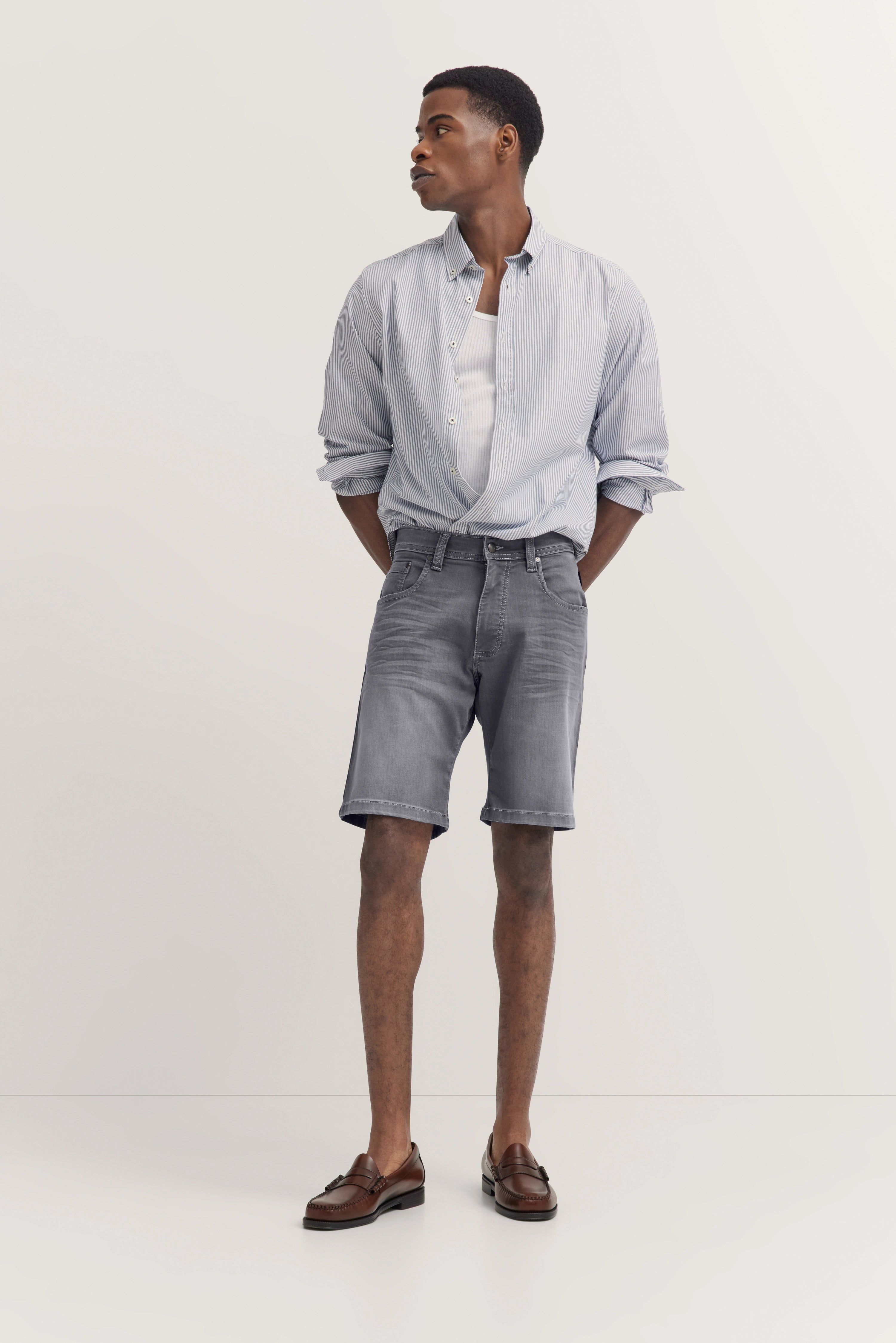 Model in gestreiftem Hemd, grauen Shorts und braunen Loafers, lässiges Posing, bugatti Stil.