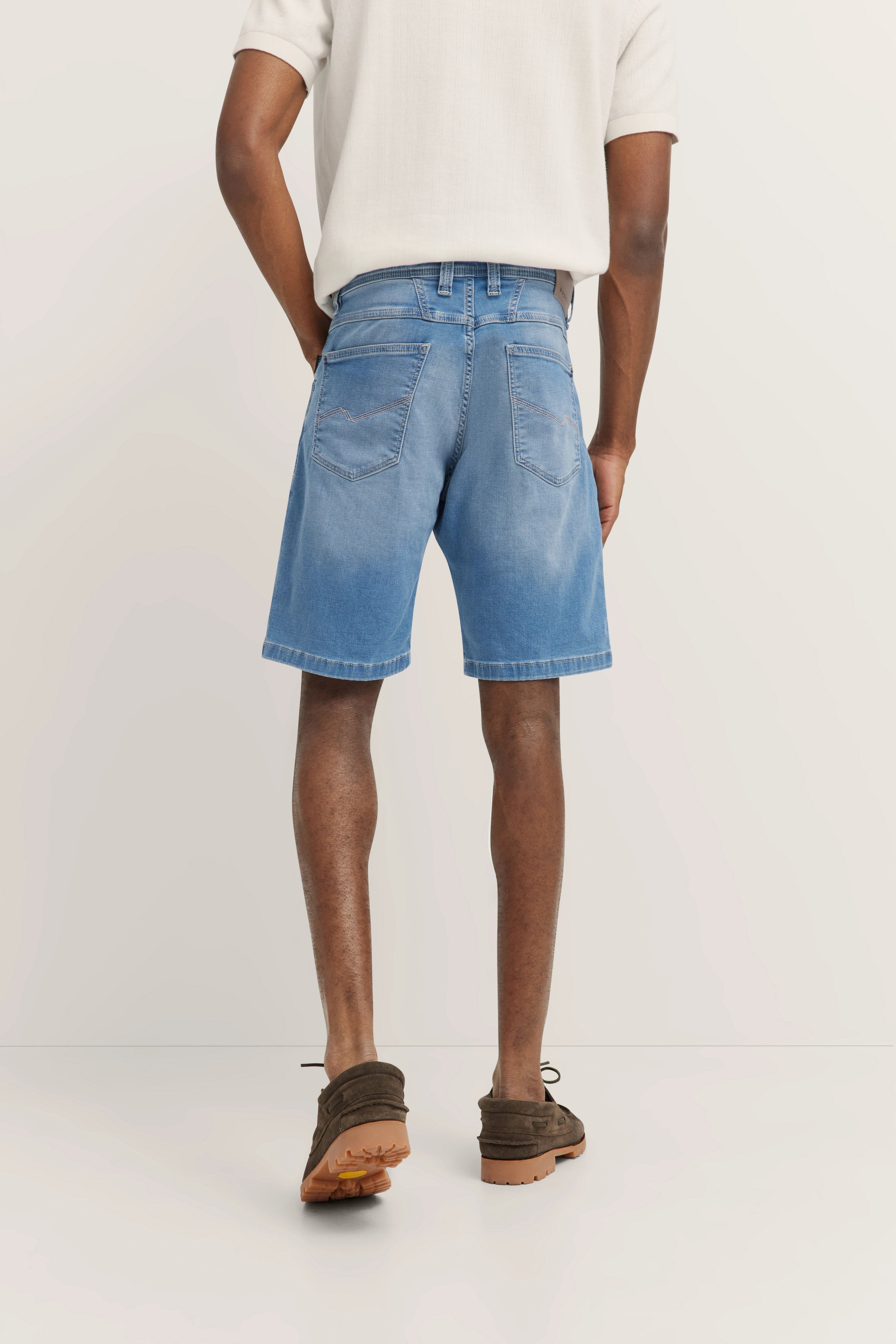bugatti Modelbild: Mann in weißen Polo, blauen Jeansshorts, braunen Schuhen, Rückenansicht, entspanntes Posing.