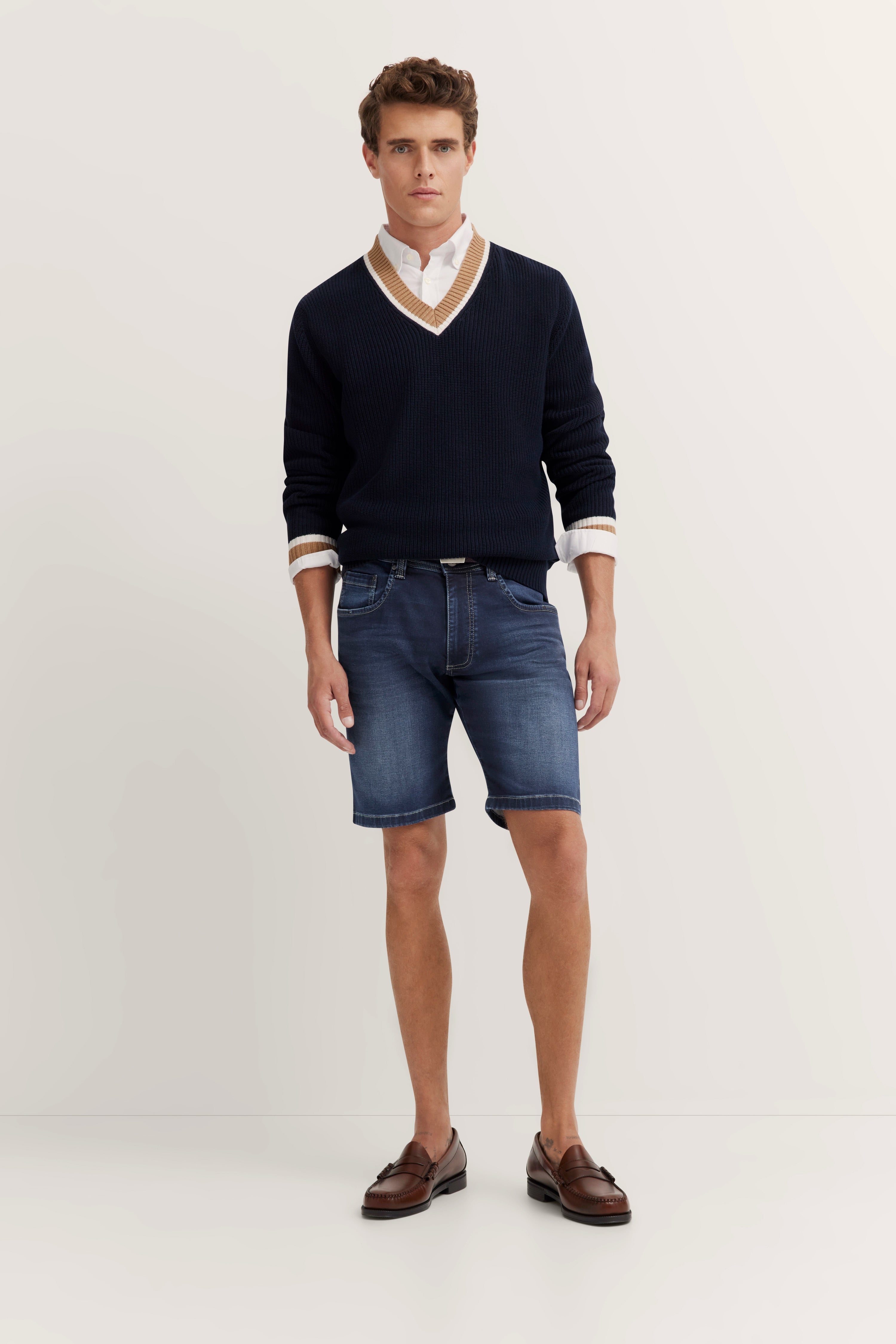 Model in bugatti Outfit: Dunkelblauer Pullover, weißes Hemd, blaue Shorts, braune Loafer. Entspanntes Posing.