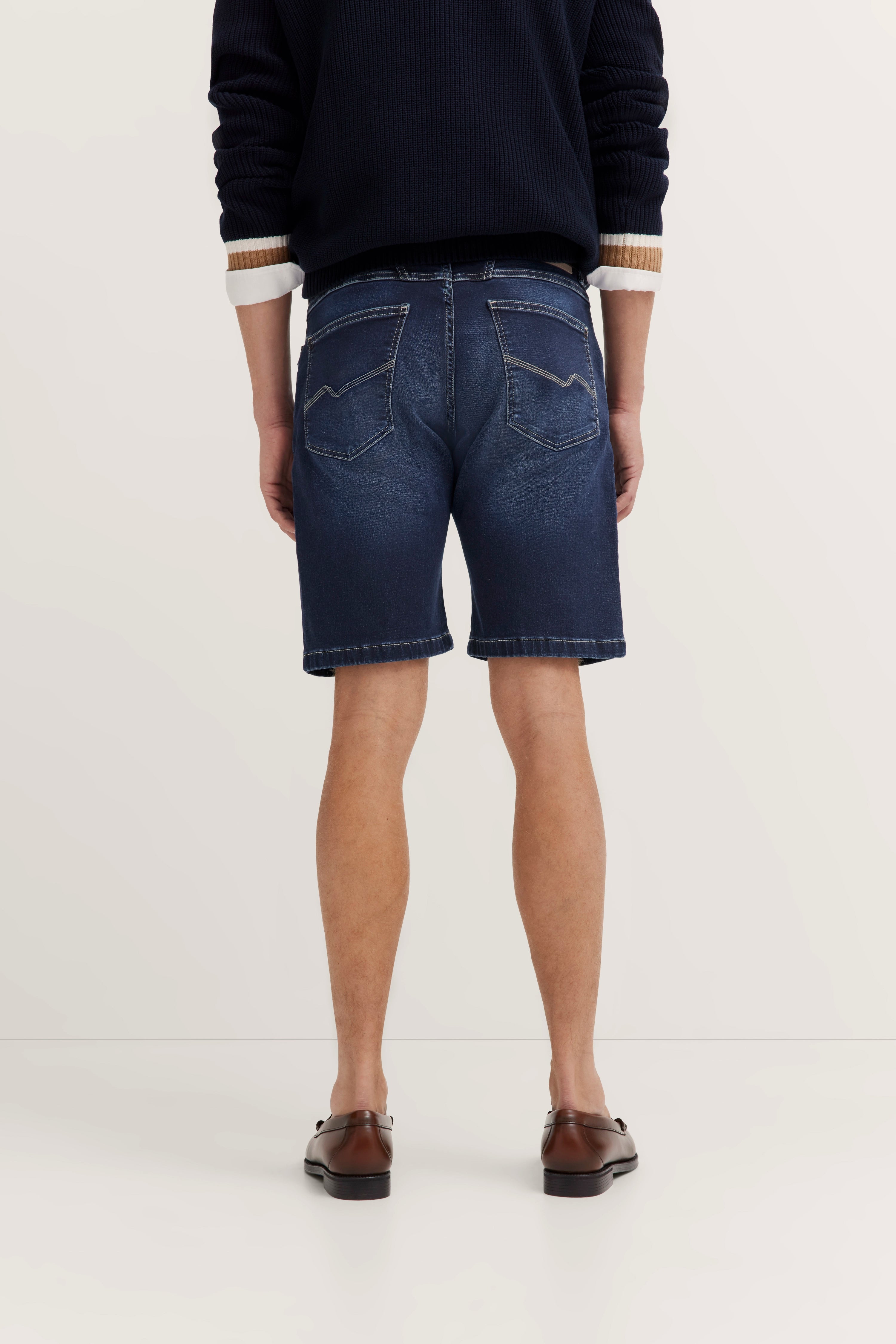 Rückansicht: Dunkelblauer Pullover, dunkelblaue Jeansshorts, braune Lederschuhe. Model stehend.