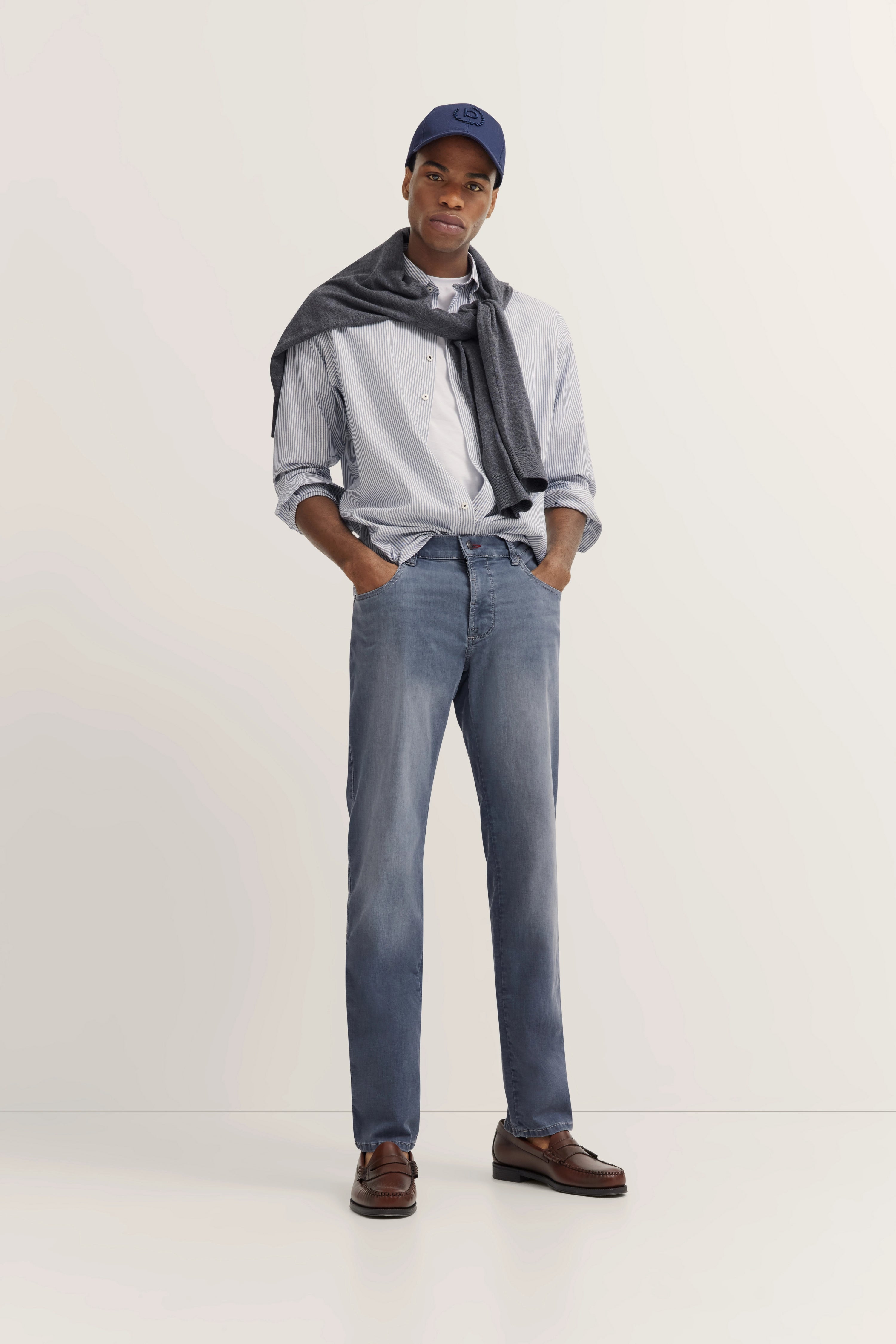 Man in hellblauem Hemd und Jeans, grauer Pullover um Schultern, blaue Kappe, braune Schuhe, lässige Pose.