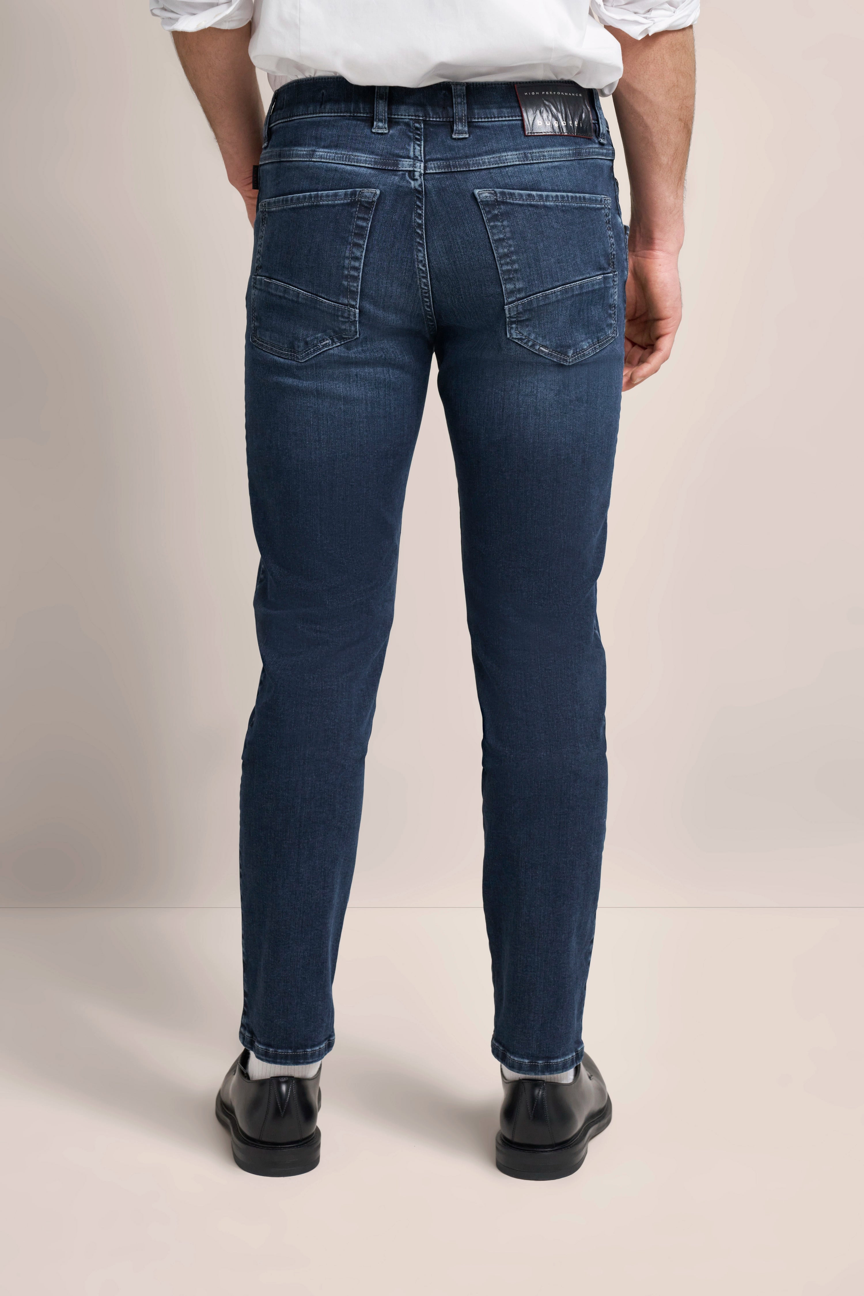 Dunkelblaue Jeans von bugatti, Rückansicht, legeres Modelbild, mit schwarzem Schuh und weißem Hemd.