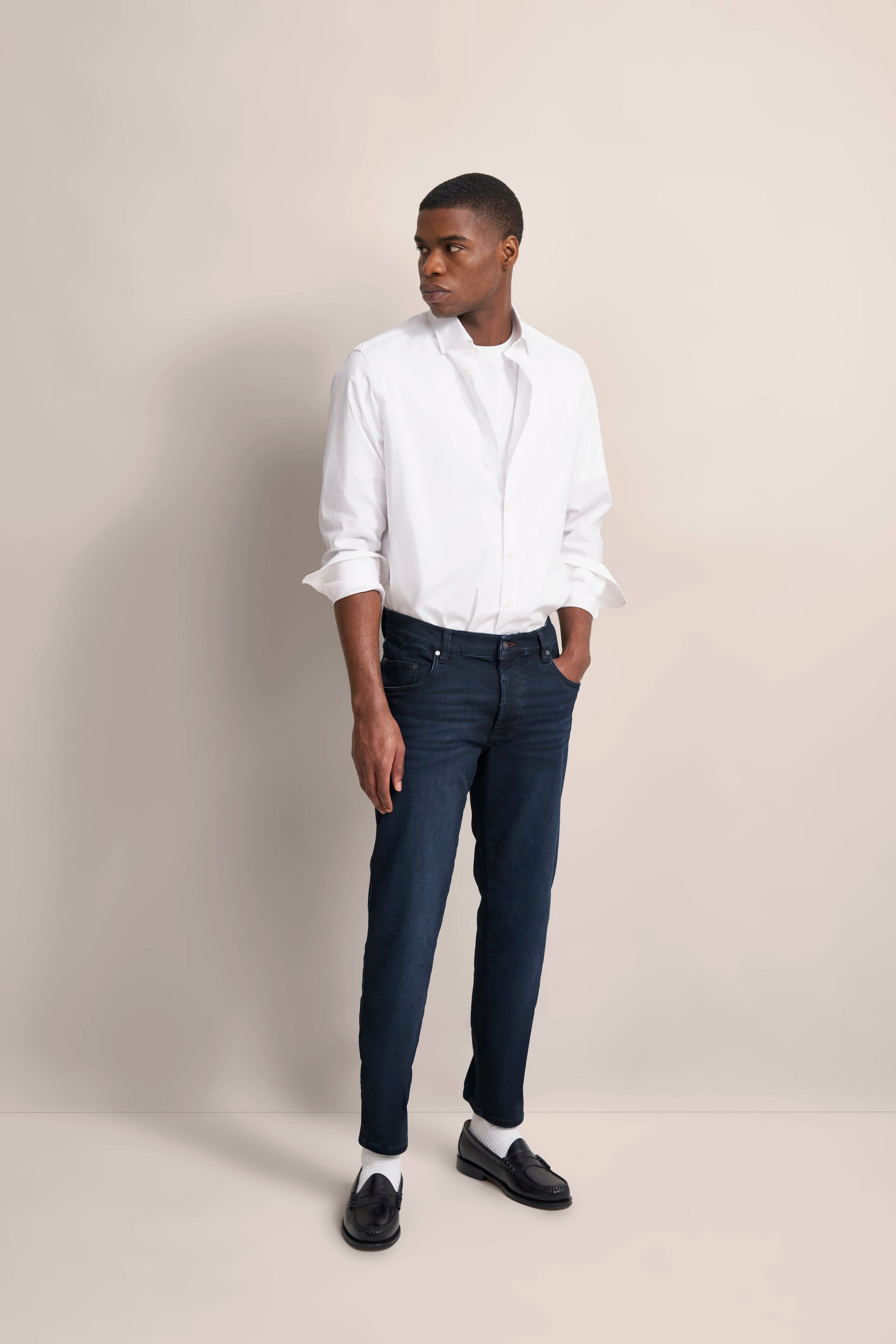 Weißes Hemd, dunkelblaue Jeans, schwarzer Loafer. Model mit Hand in Tasche, seitlicher Blick.