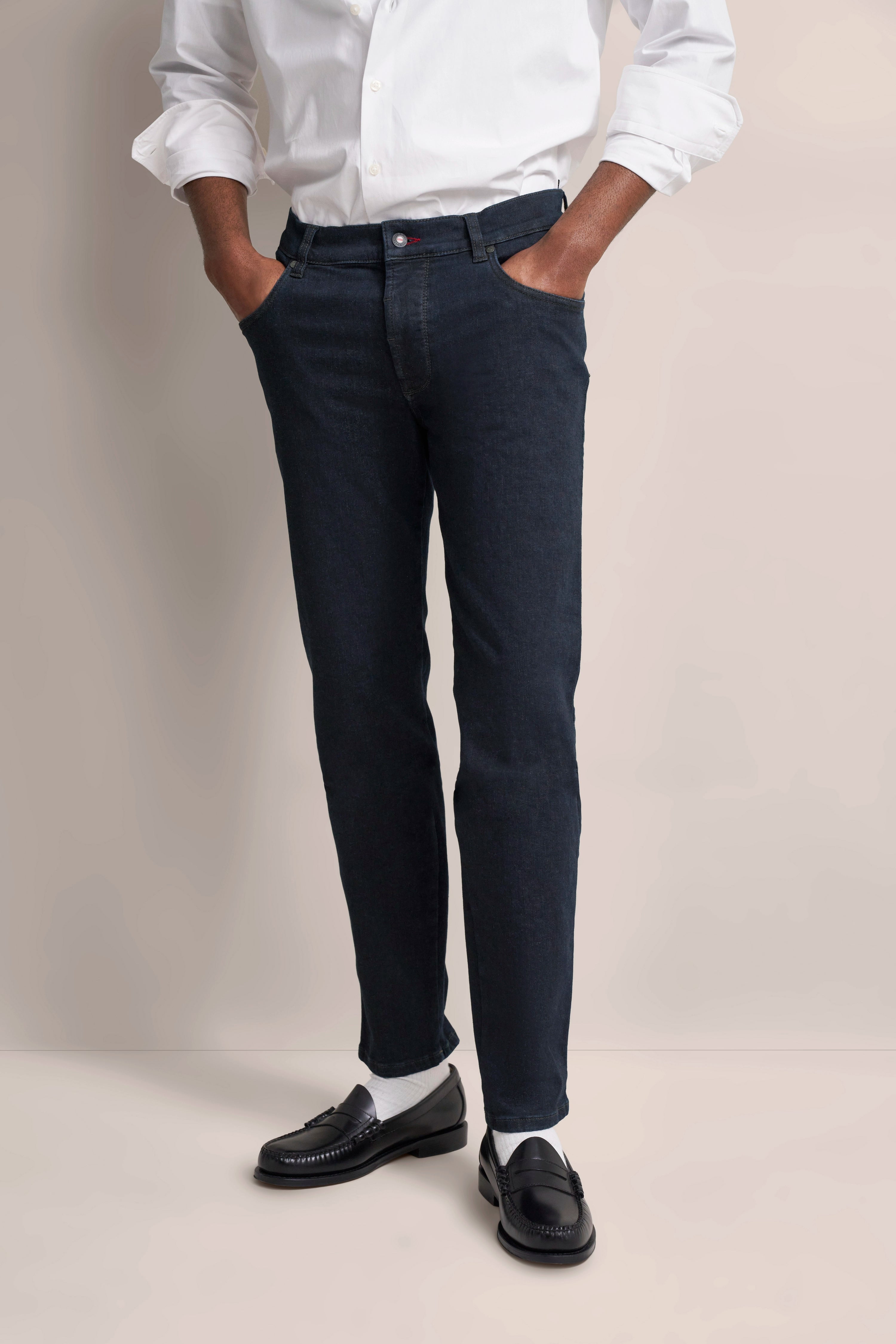 bugatti Modelbild: Weißes Hemd, dunkelblaue Jeans, schwarze Loafer. Hände in Hosentaschen, lässige Pose.