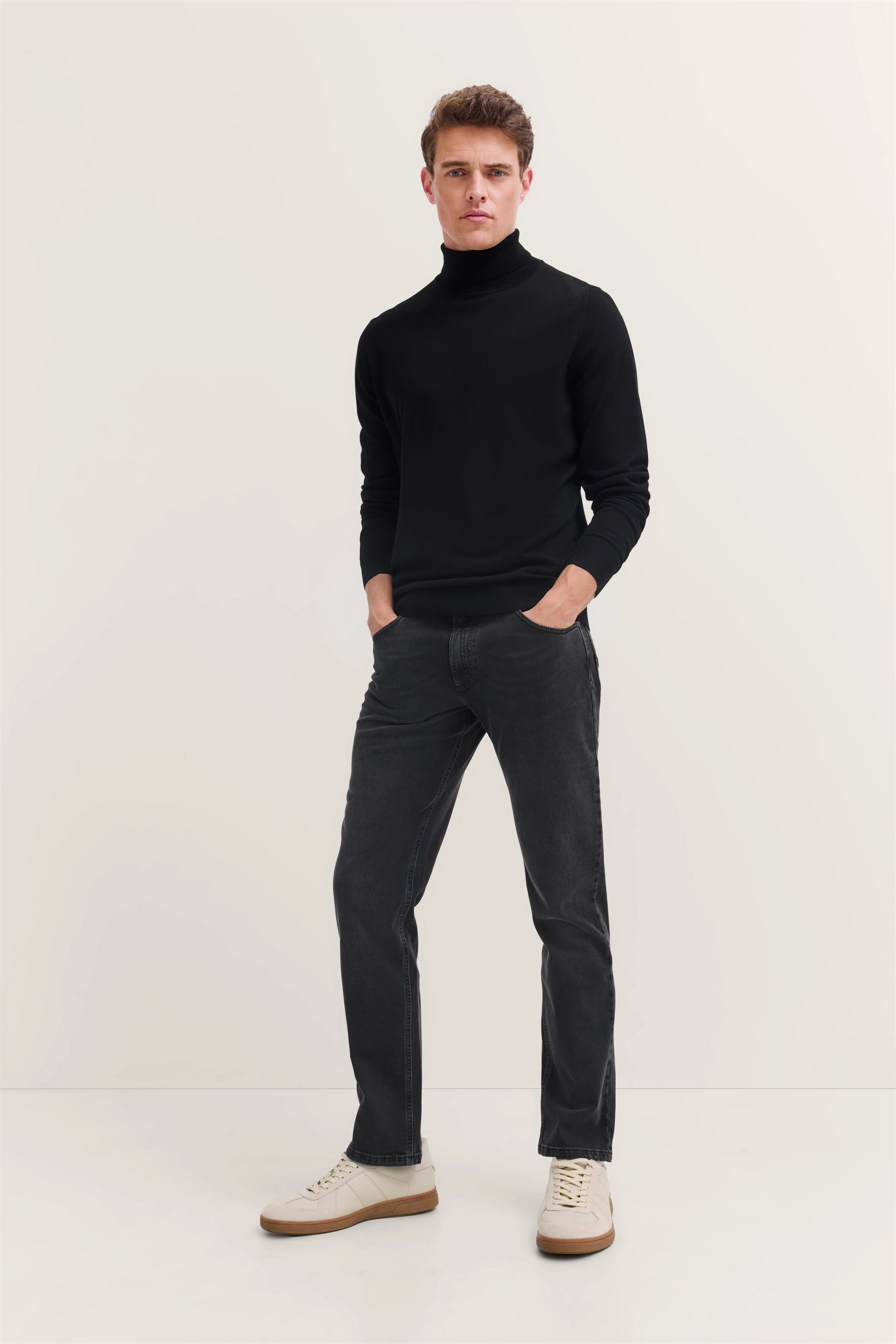 Model in schwarzem Rollkragenpullover und grauer Jeans, lässig stehend, helle Sneaker.