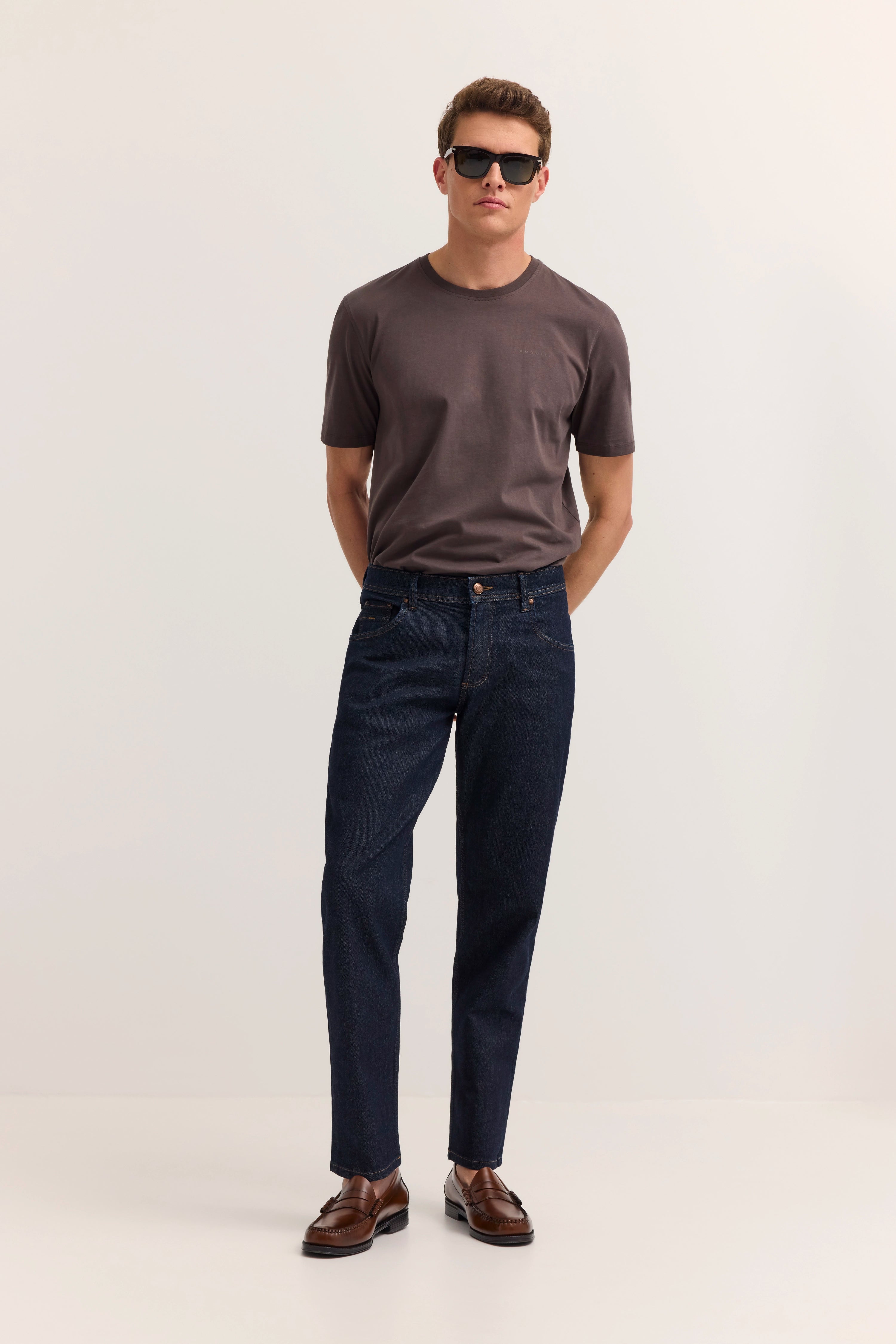 bugatti Model in grauem T-Shirt und dunkler Jeans, lässige Pose, Hände in Hosentaschen.