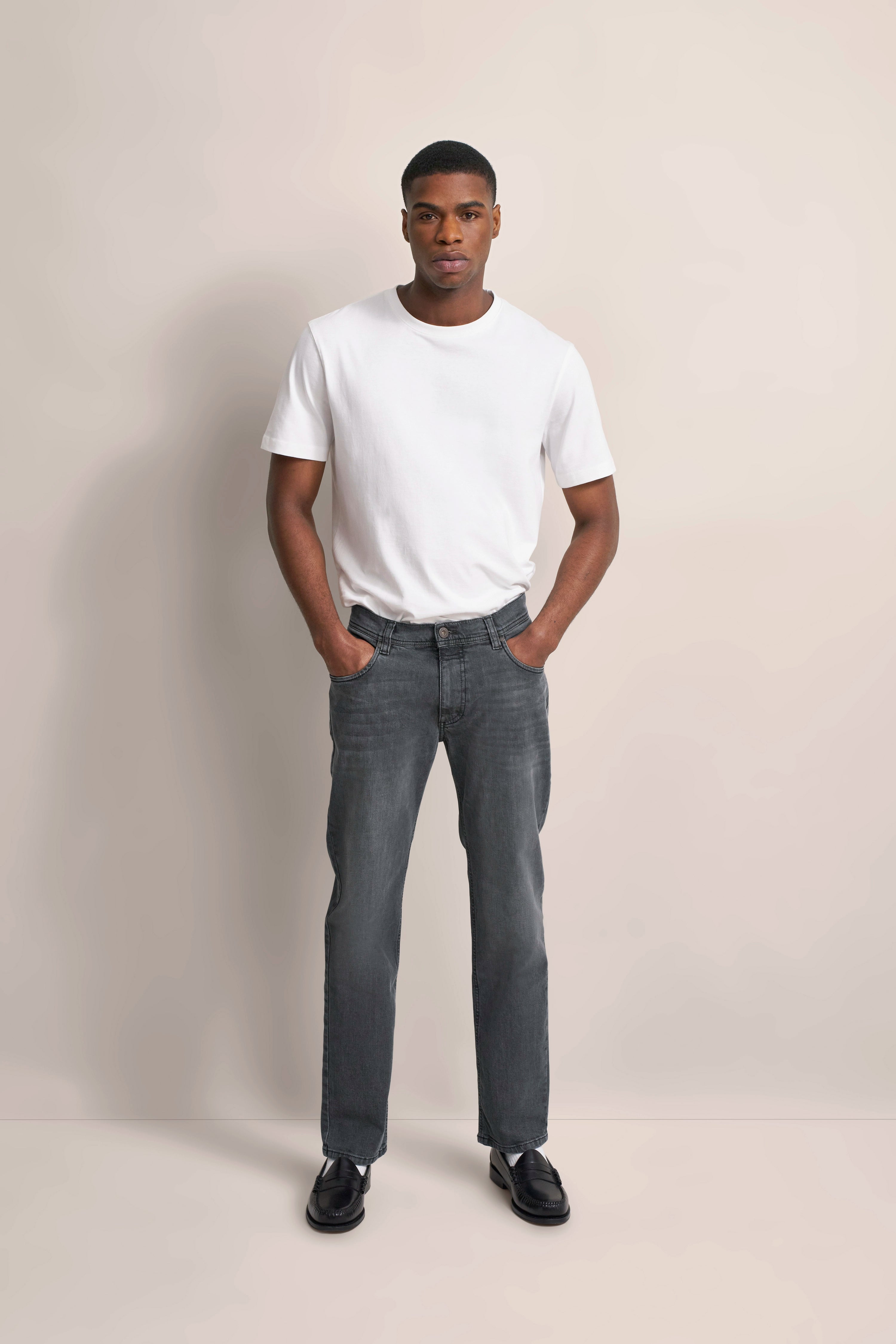 Modelbild: Weißes T-Shirt, graue Jeans, schwarze Schuhe, lässige Pose, Hände in den Hosentaschen.