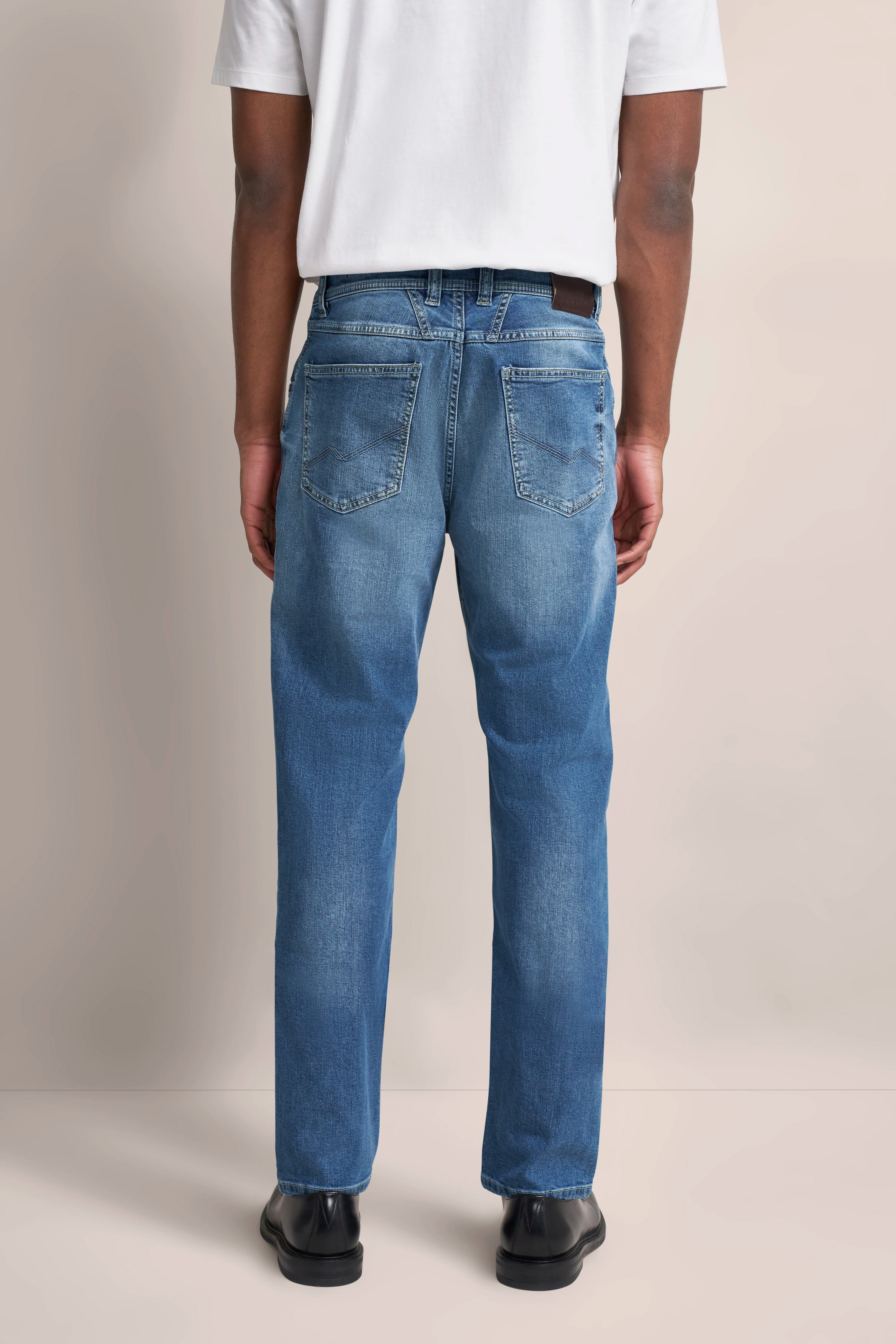 Blau Jeans rückseitig, legeres Modelbild. Model trägt weißes Shirt und schwarze Schuhe.