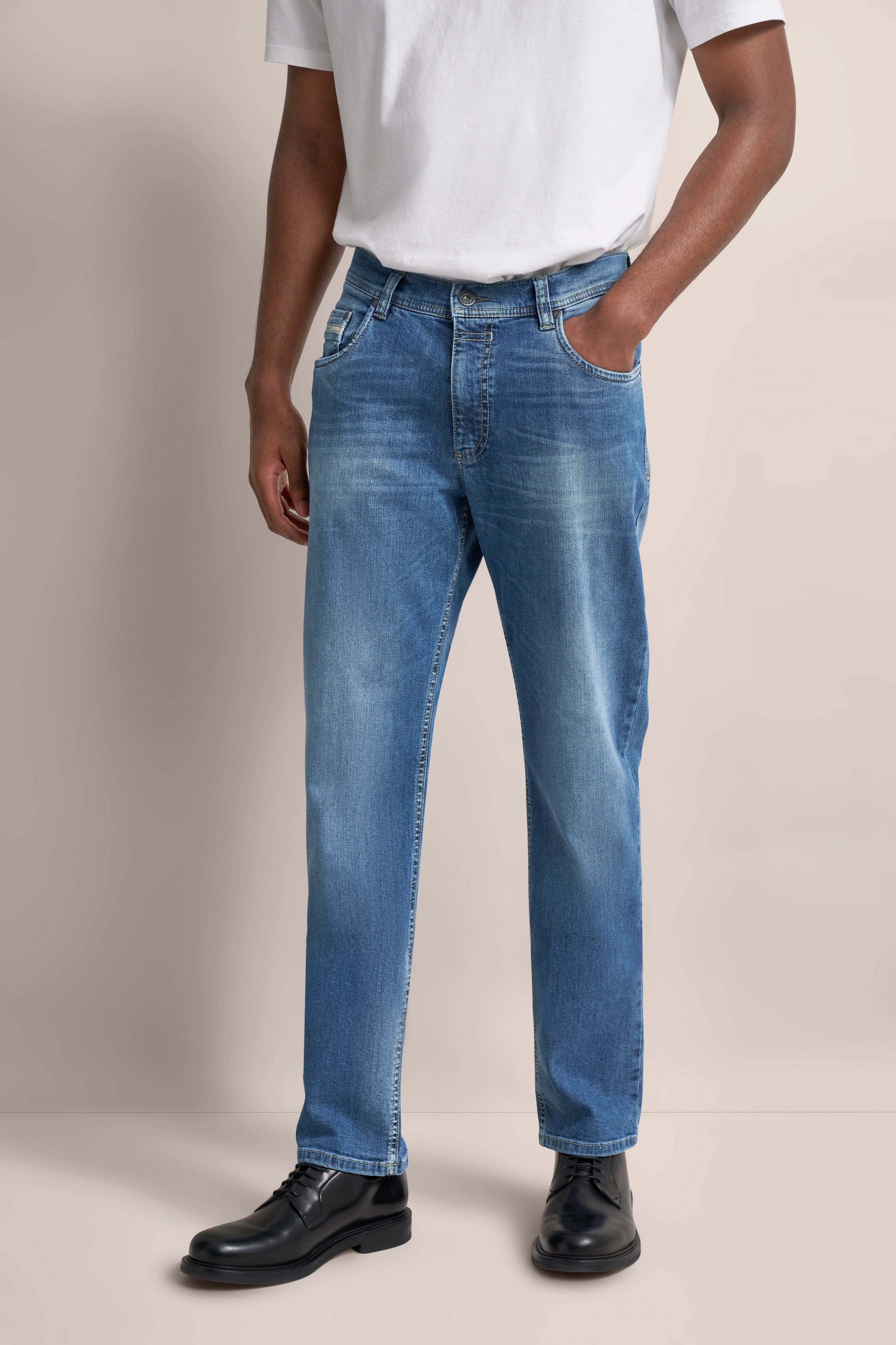 Jeans in Blau mit weißem T-Shirt, schwarzer Schuh. Model stehend, Hand in Hosentasche.