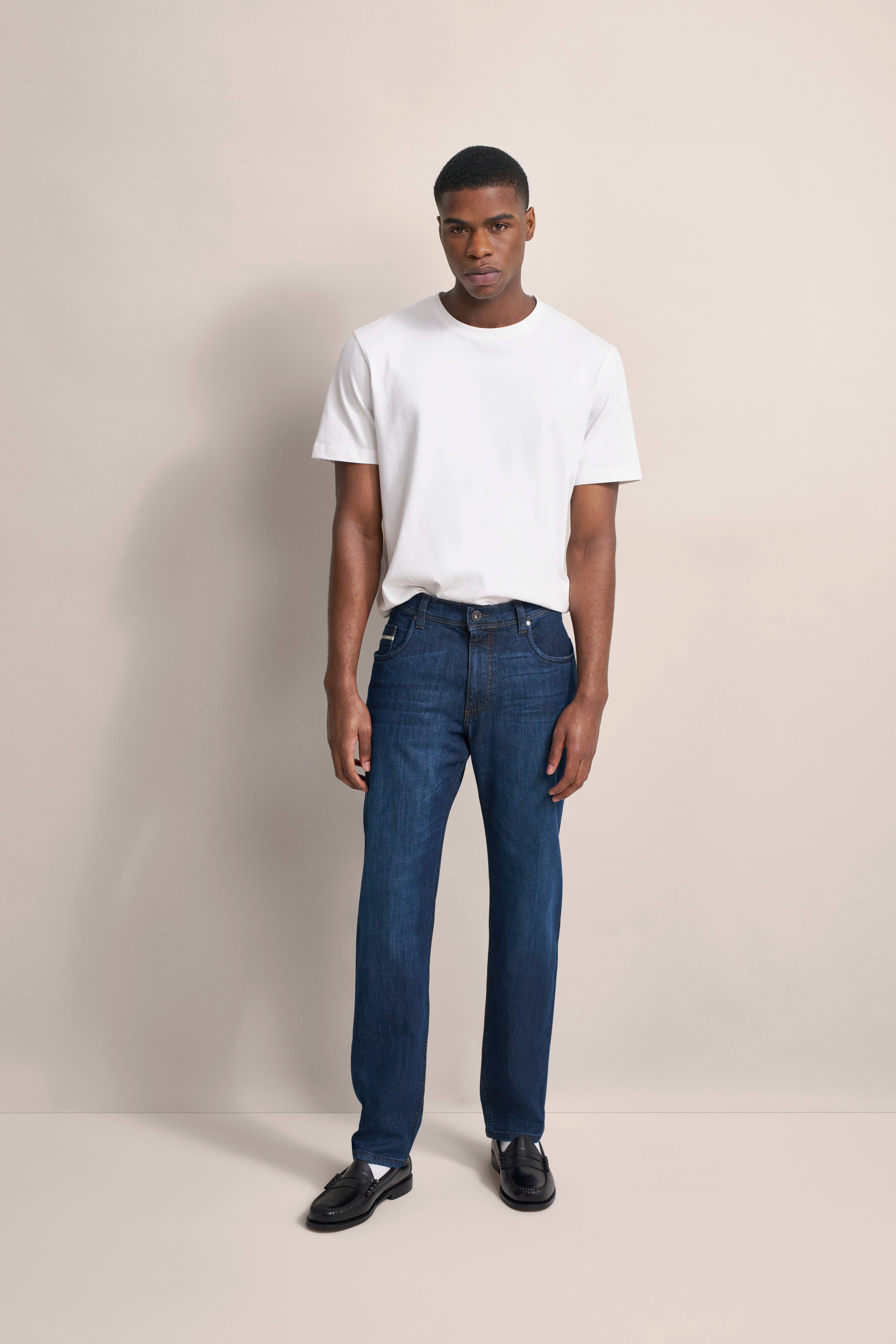 Model in weißem T-Shirt und blauer Jeans, steht entspannt. Outfit von bugatti.
