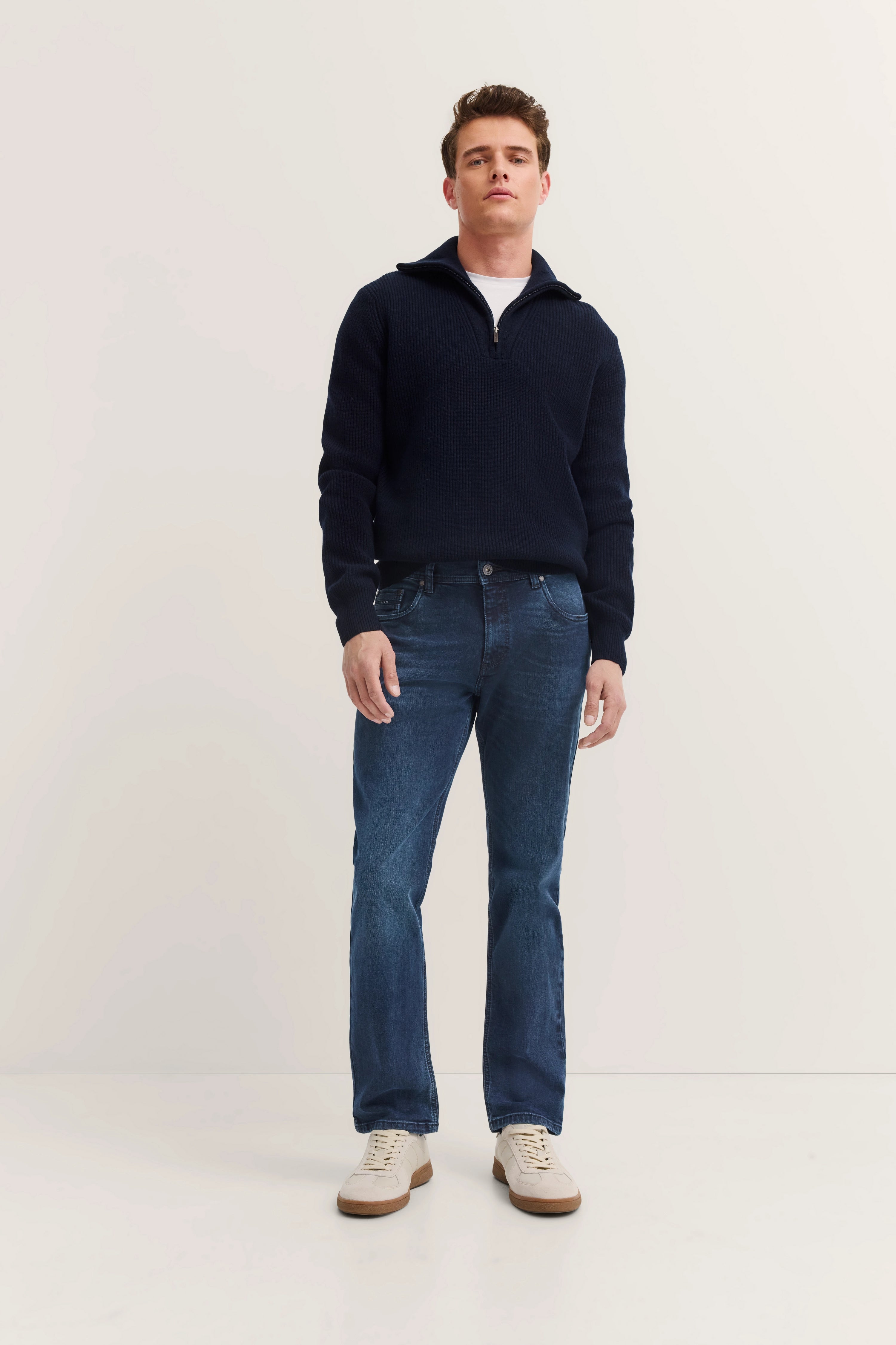 Modelbild: Mann in dunkelblauem Pullover, blauen Jeans, weißen Sneakern, steht frontal, entspanntes Posing.