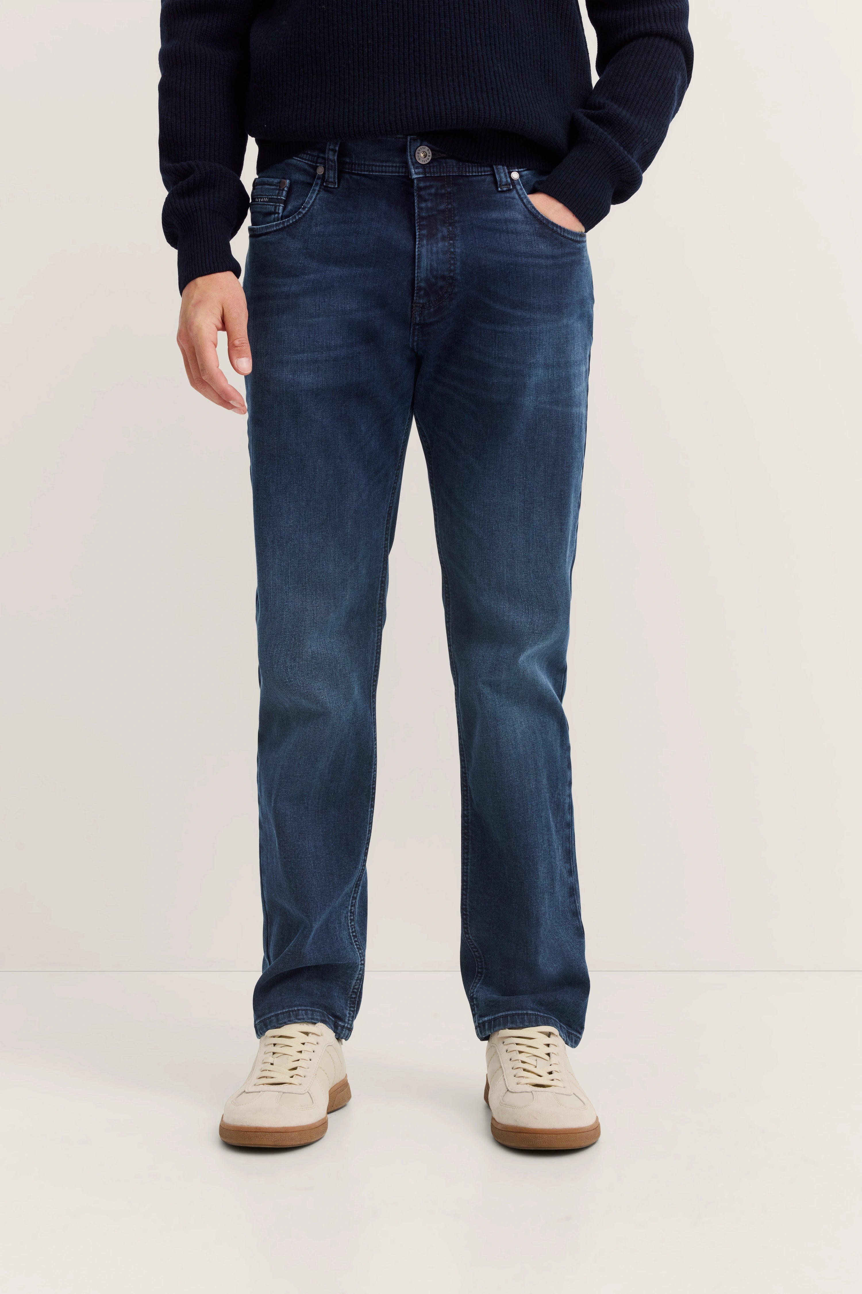 Dunkelblaue Jeans mit schwarzem Pullover, legeres Modelbild, Hände in Taschen, weiße Sneaker.