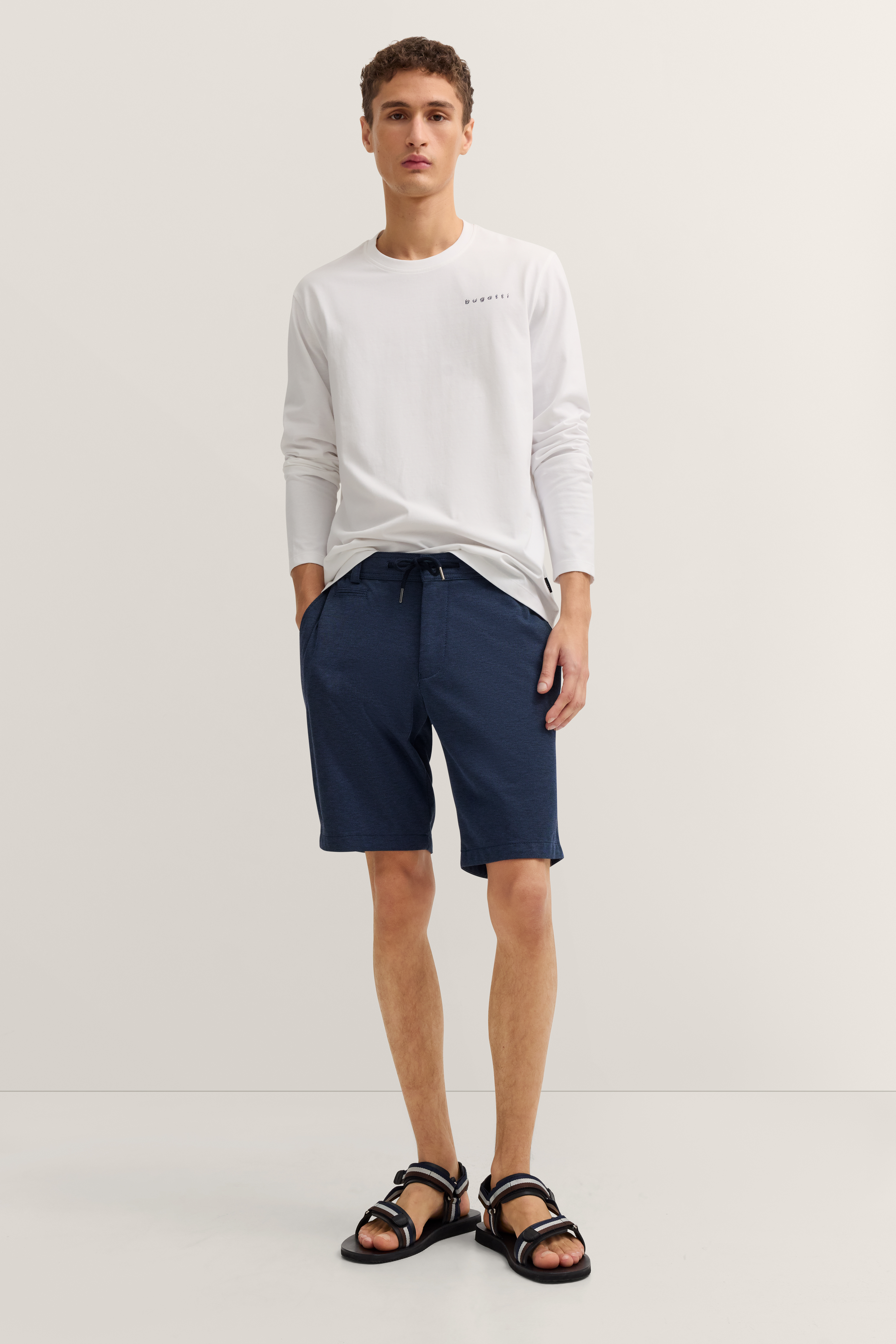 Weißes bugatti Langarmshirt, blaue Shorts, schwarzes Sandalen-Model, lässige Pose.