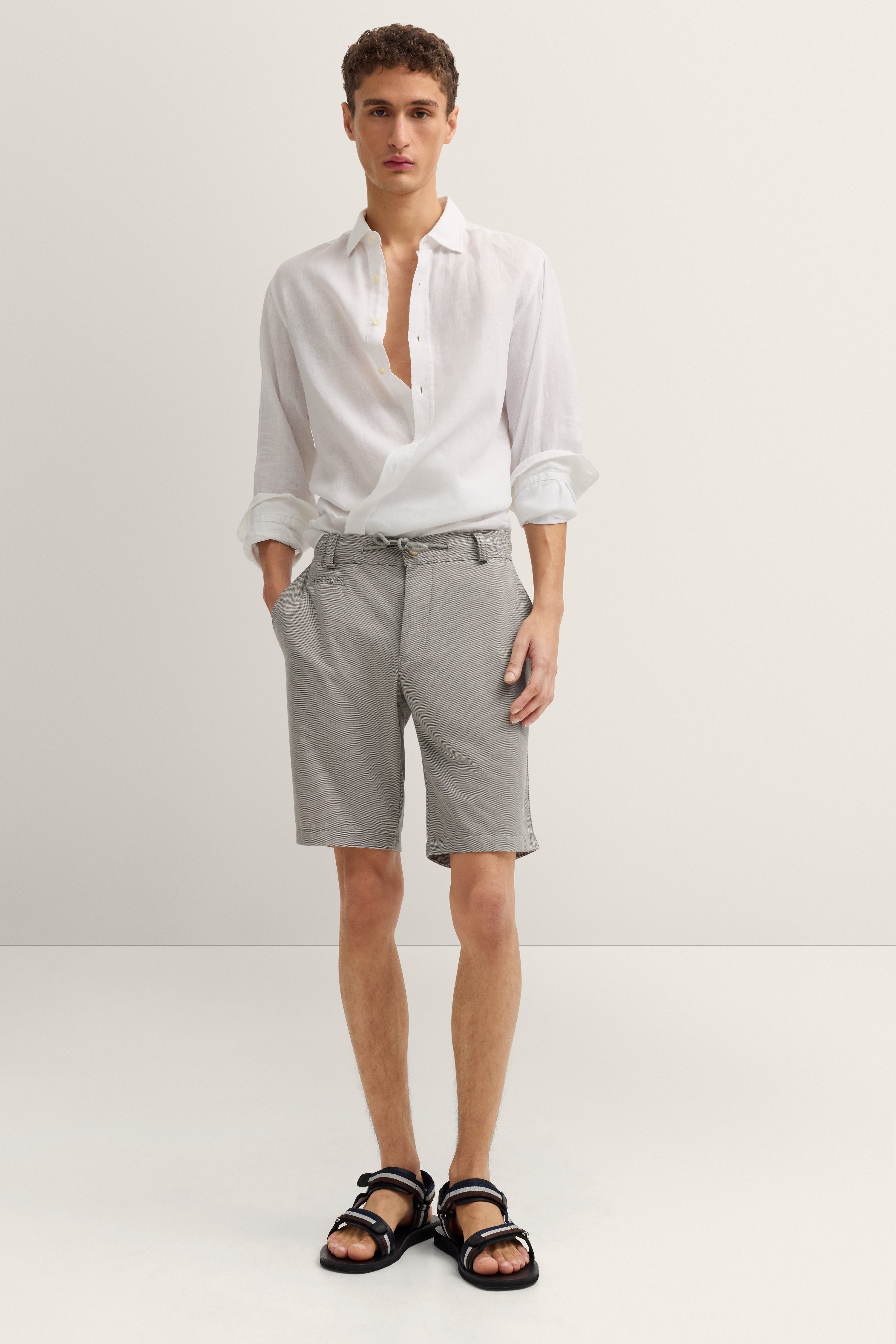 Rückansicht eines legeren Outfits: graue Shorts, weißes Hemd, schwarze Sandalen.