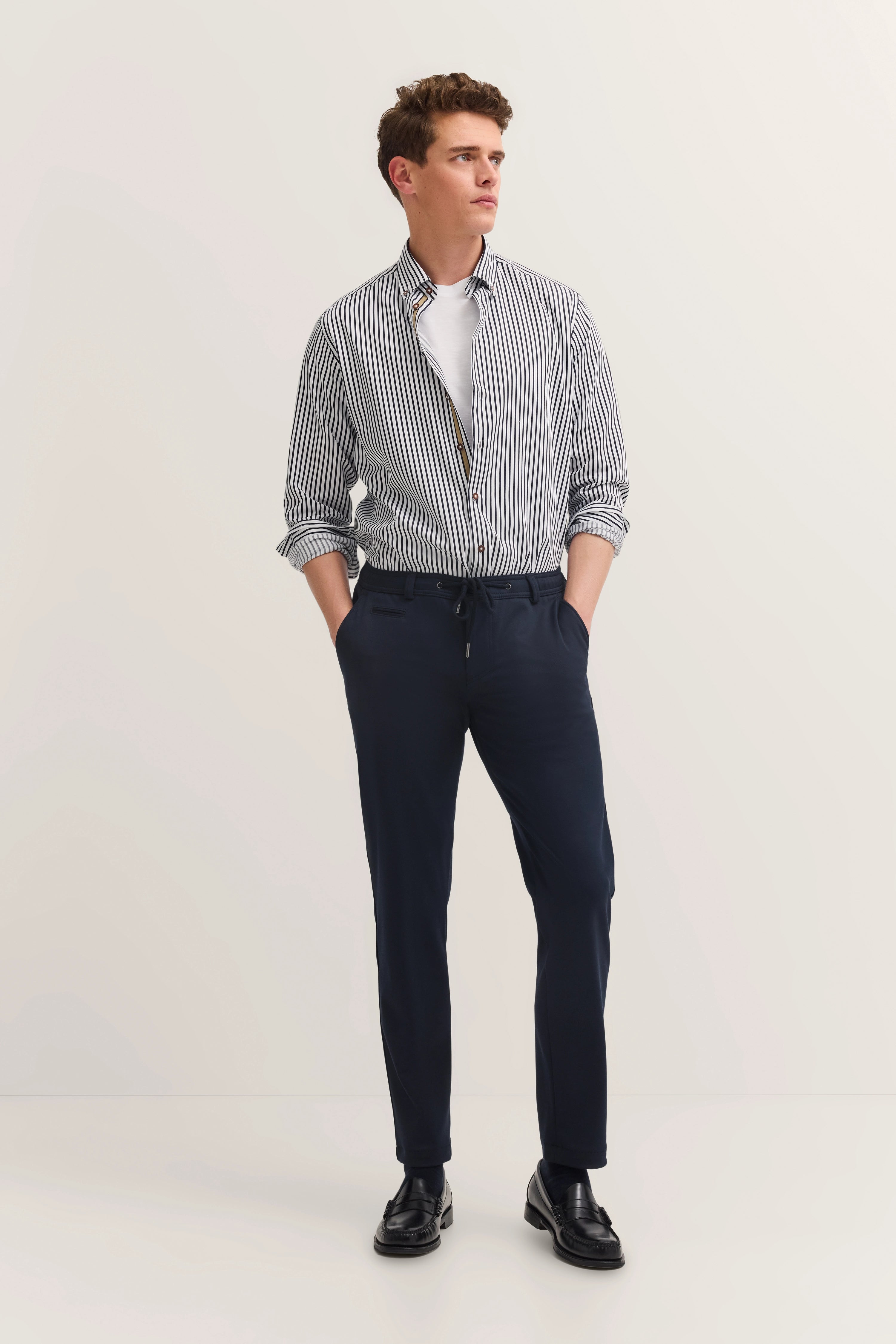 Model im gestreiften Hemd und blauer Hose, Hände in Hosentaschen, seitlich schauend.
