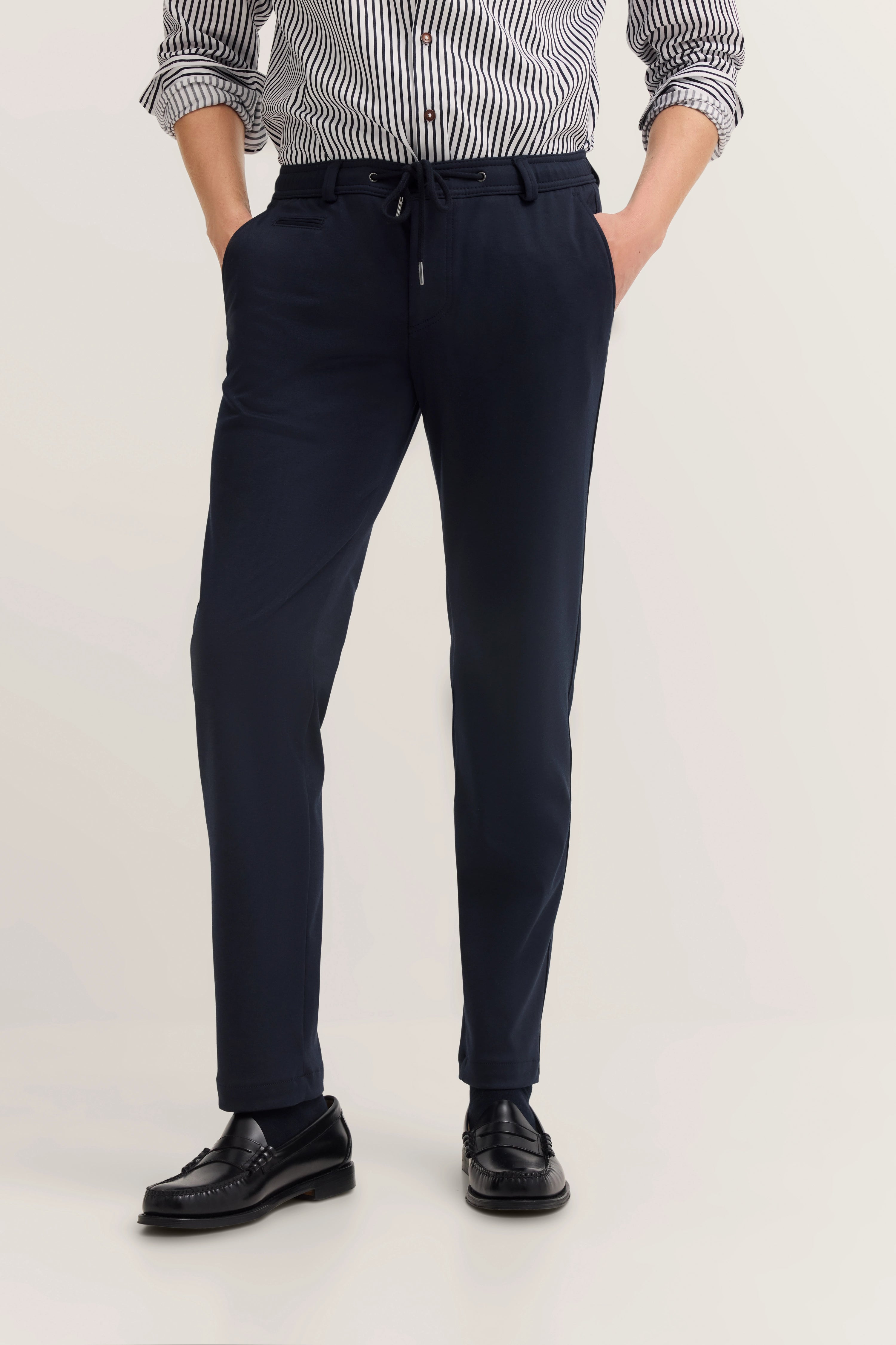 Bugatti Herren-Outfit mit gestreiftem Hemd, dunkelblauer Hose, schwarzen Loafern. Legerbild.