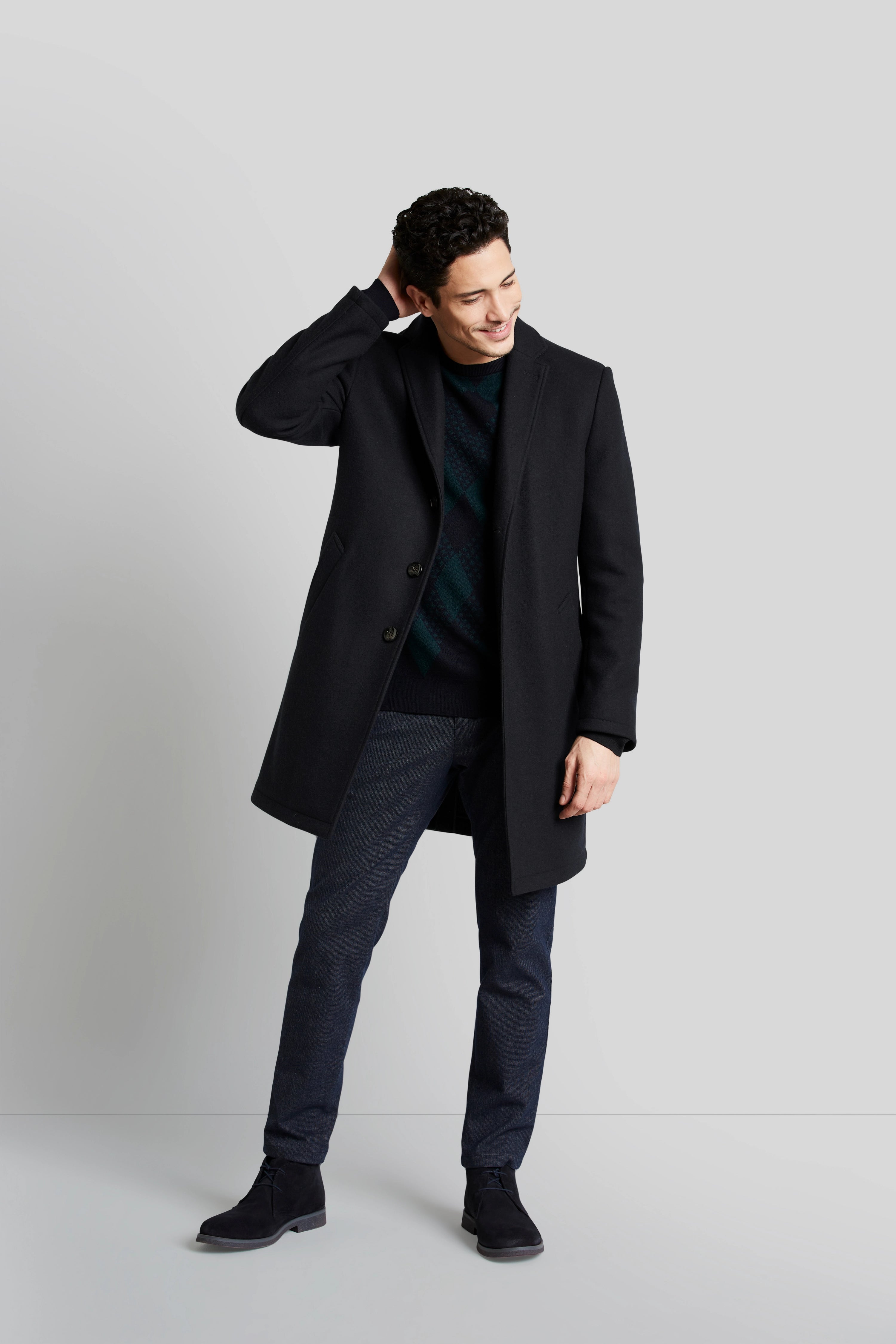 Model in schwarzem bugatti-Mantel, kariertem Pullover, blauen Hosen, lässig stehend, Hand am Kopf.