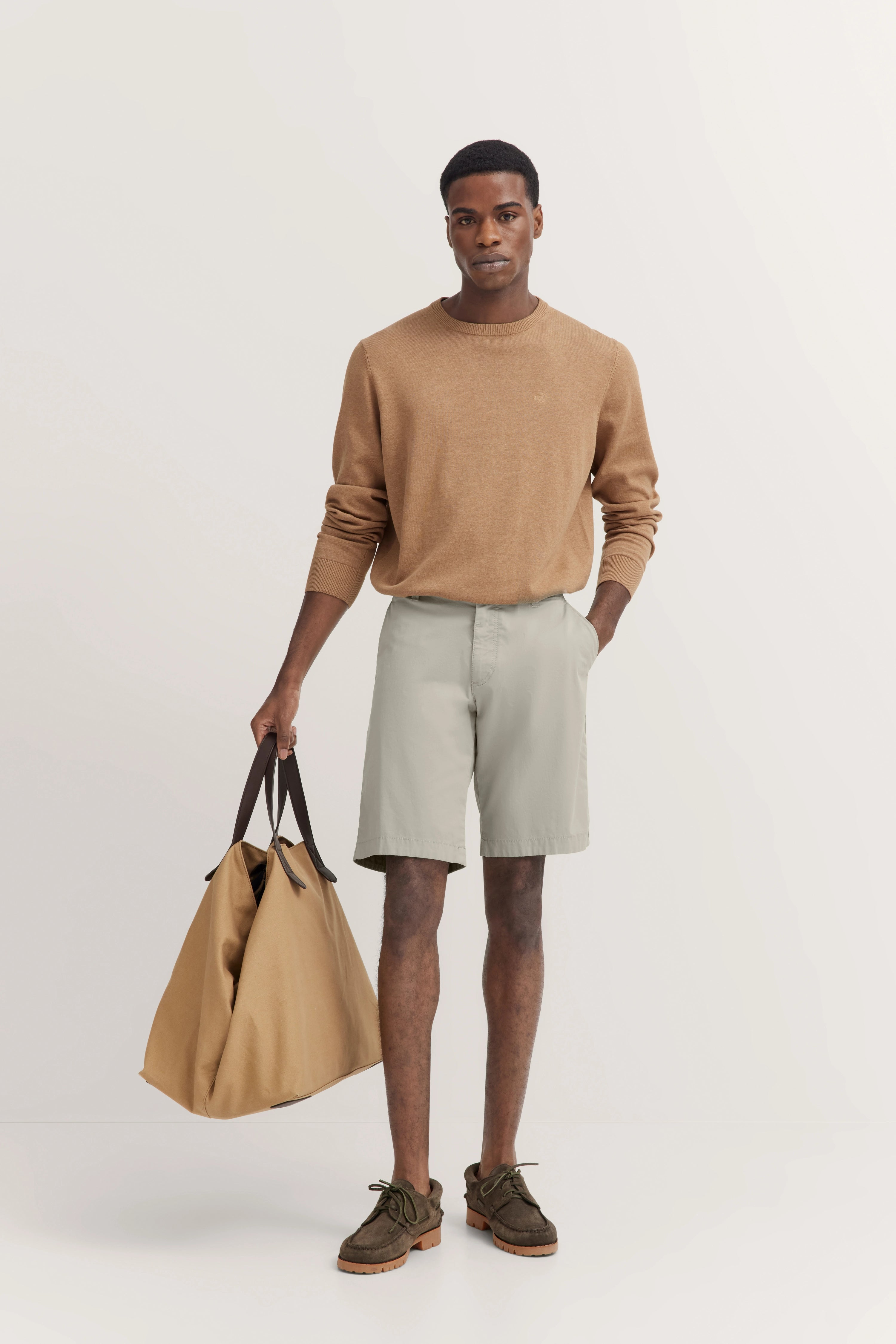 Model in beigem Pullover, hellen Shorts, mit großer Tasche. Lässige Pose, bugatti Stil.