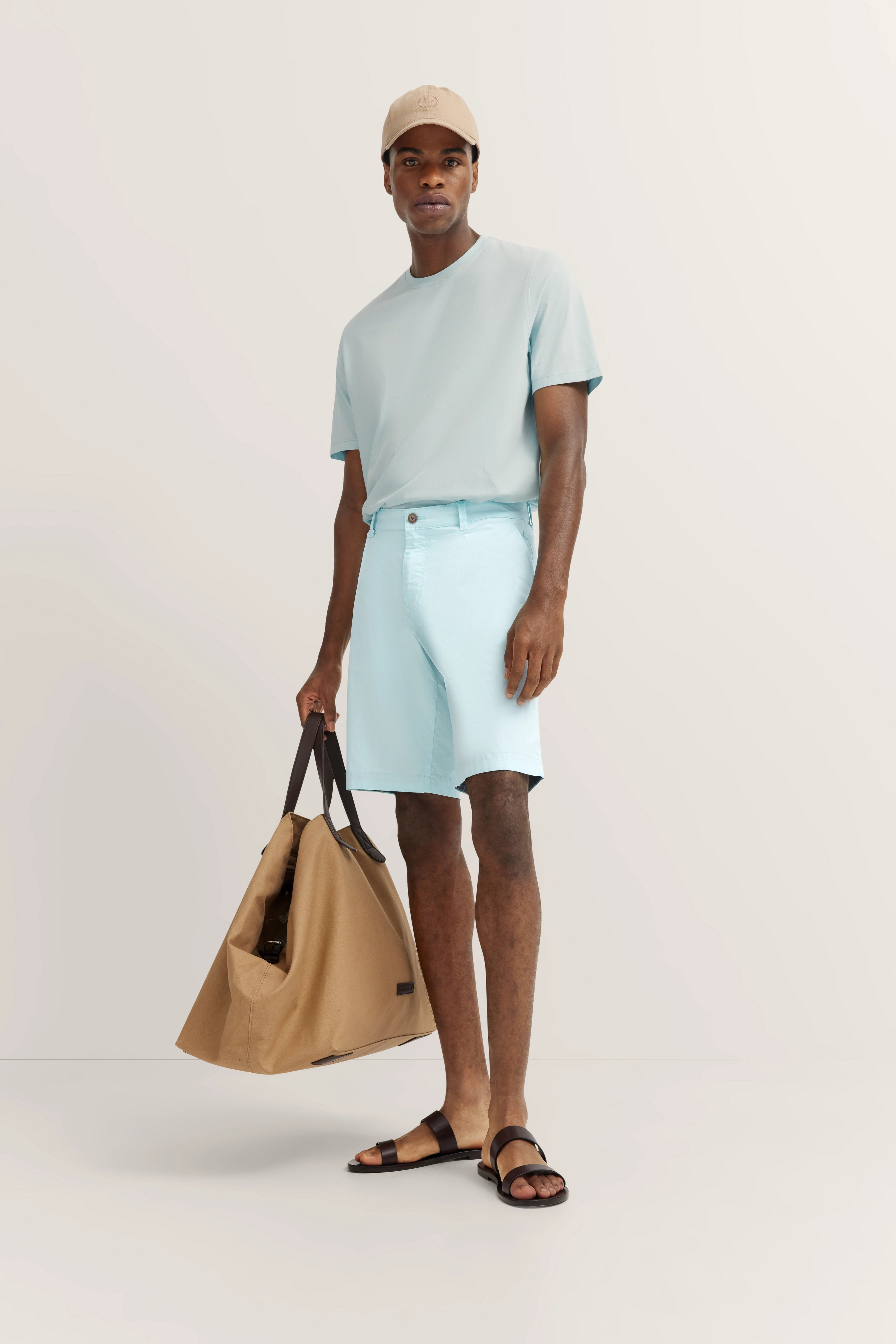 Model in hellblauem T-Shirt und Shorts, beige Kappe, braune Sandalen, sandfarbene Tasche, lässige Pose.
