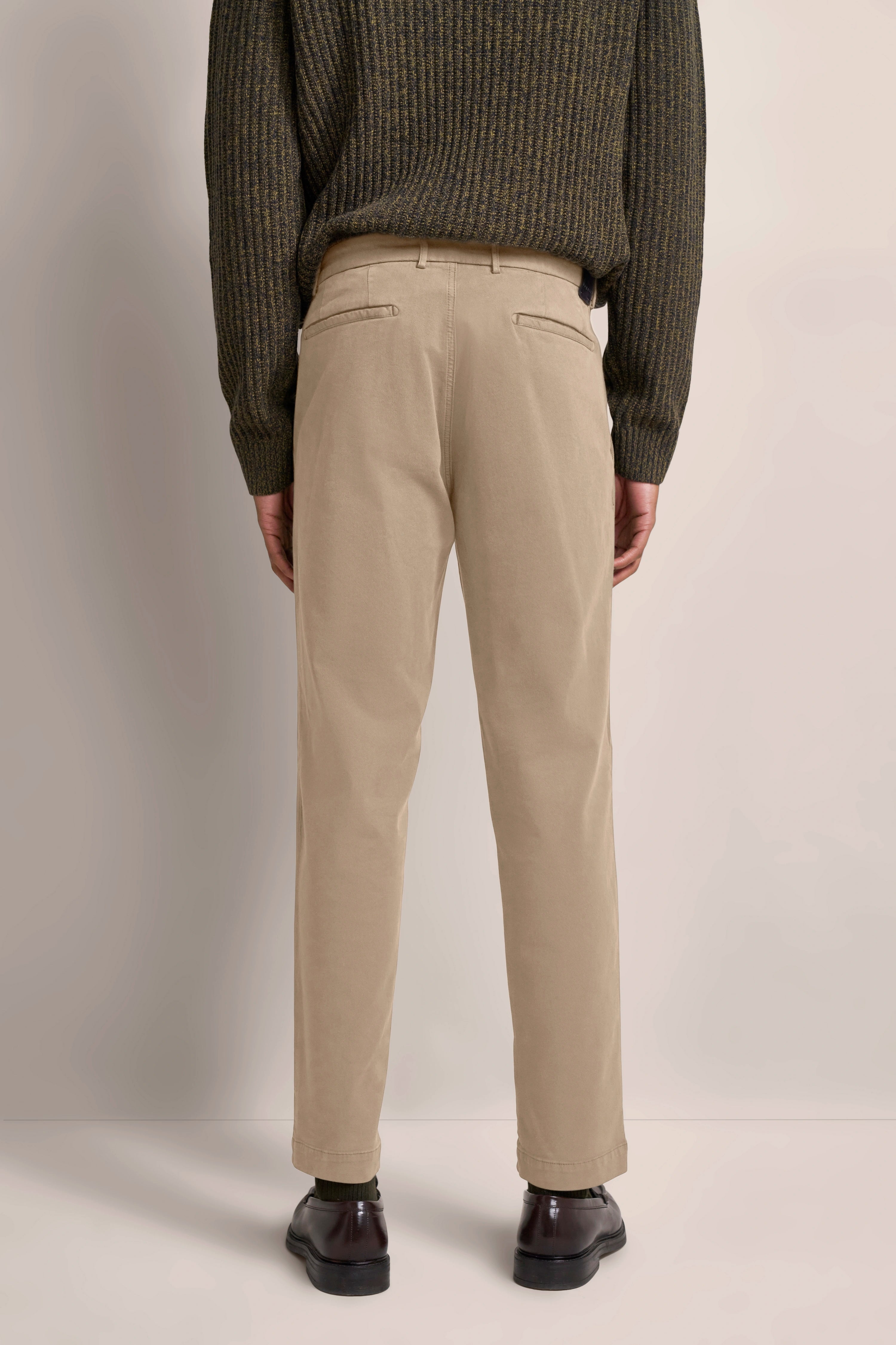 Beiger Chino mit dunkelgrünem Pullover, Model von hinten, entspannt stehend.