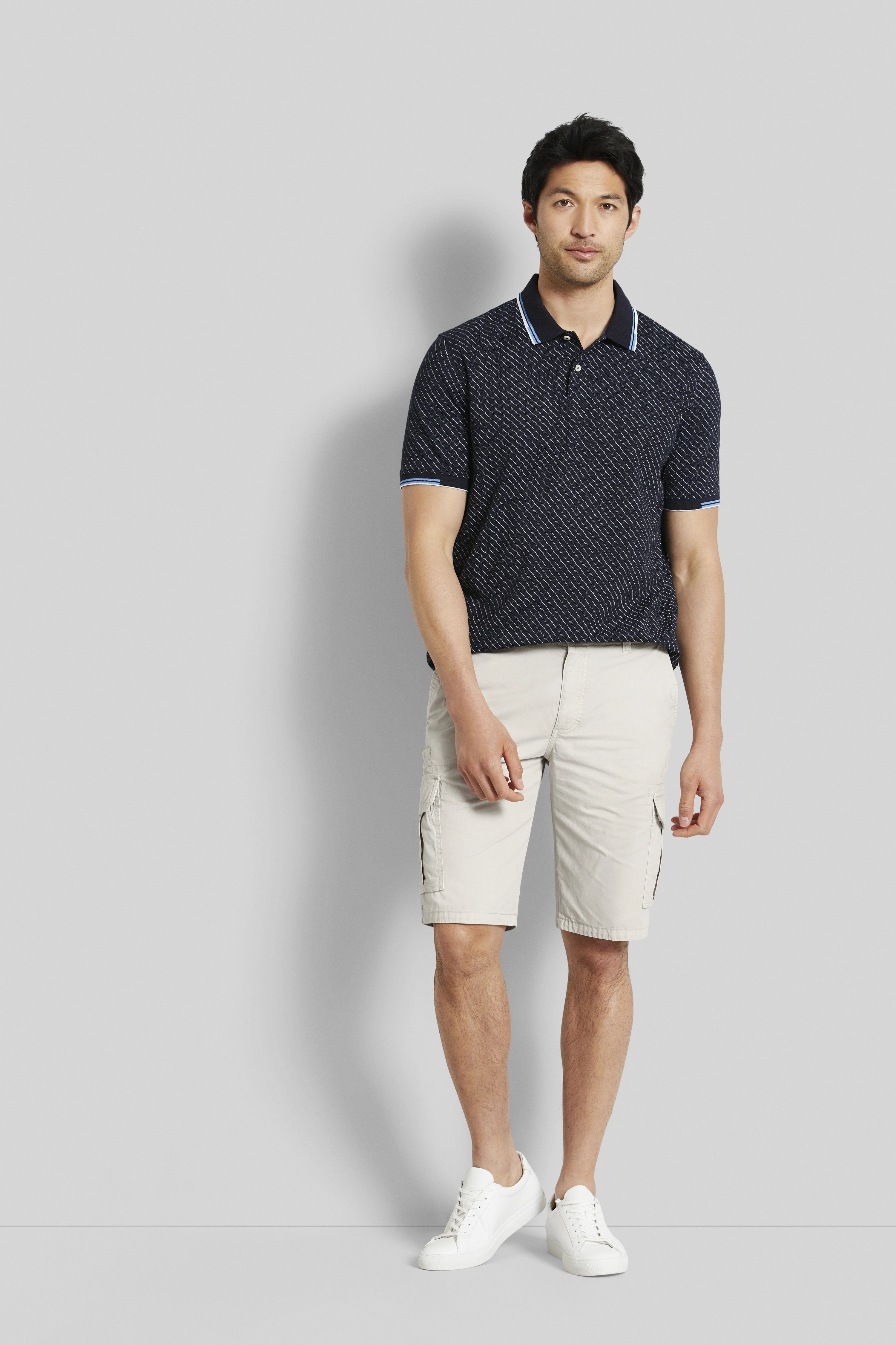Model in bugatti dark blue patterned polo, beige shorts, white sneakers, relaxed pose.