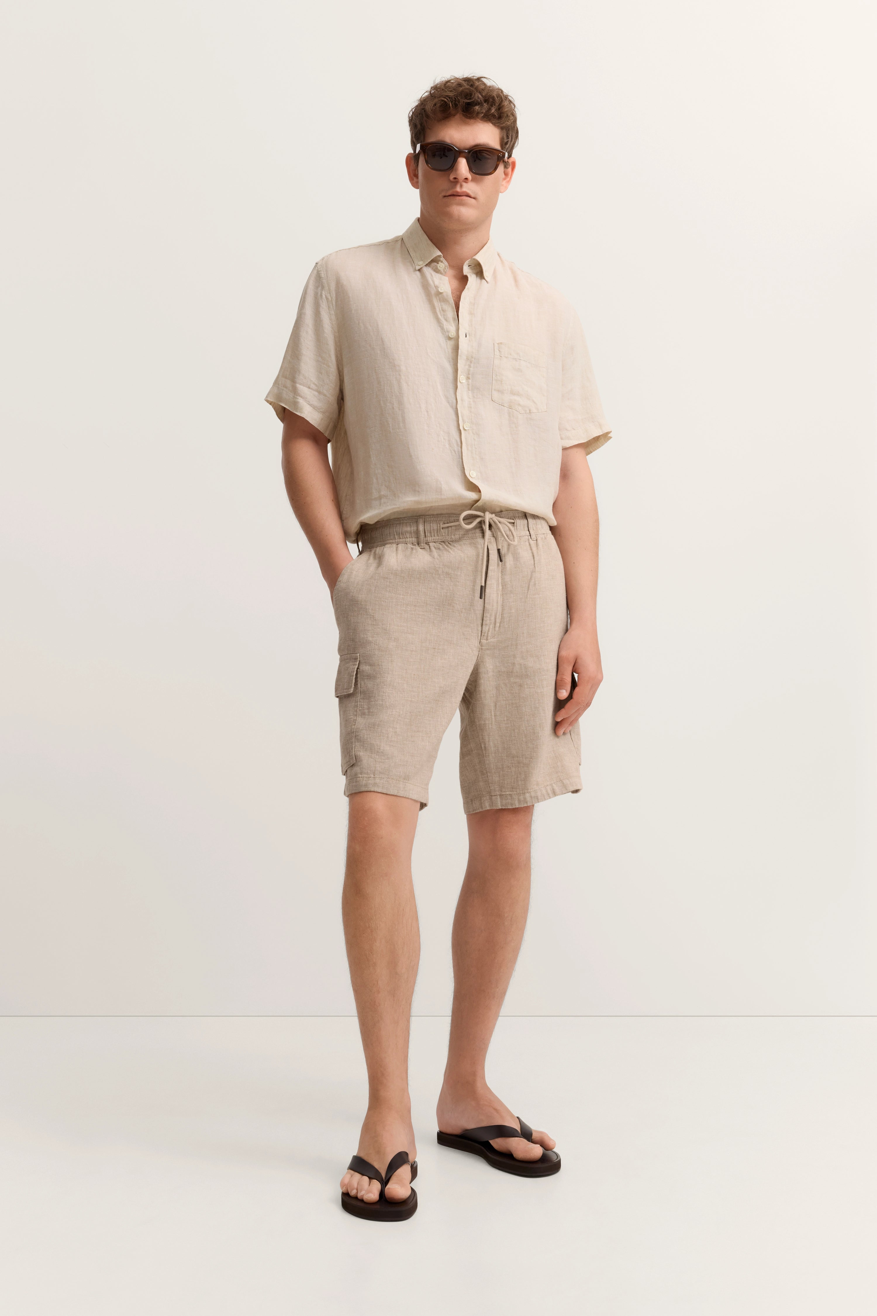 Beiges Freizeitoutfit mit Hemd und Shorts, Model posiert lässig, trägt Sonnenbrille.