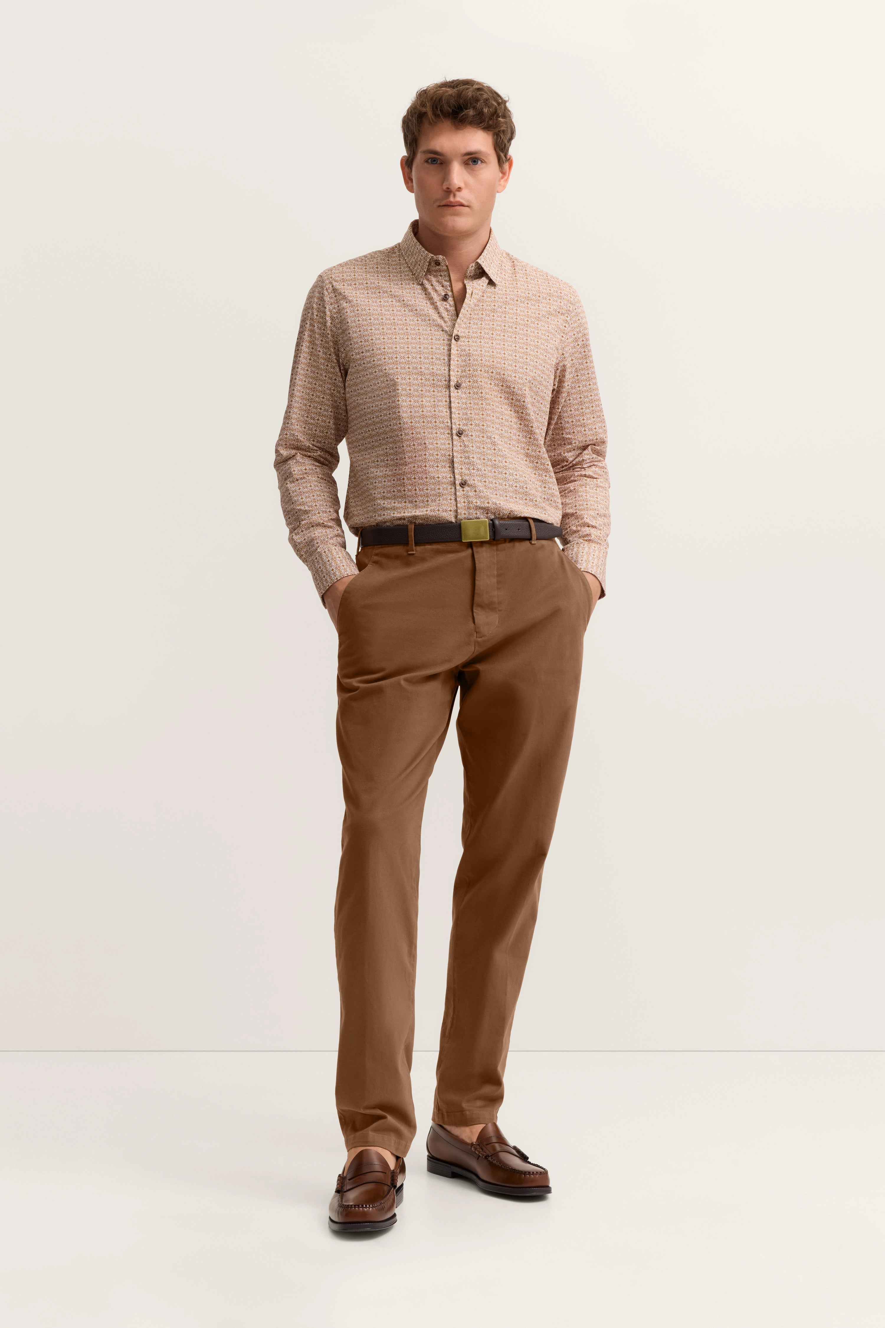 bugatti Modelbild: Beige gemustertes Hemd, braune Chinohose, braune Loafer. Hände in Taschen, entspannte Haltung.