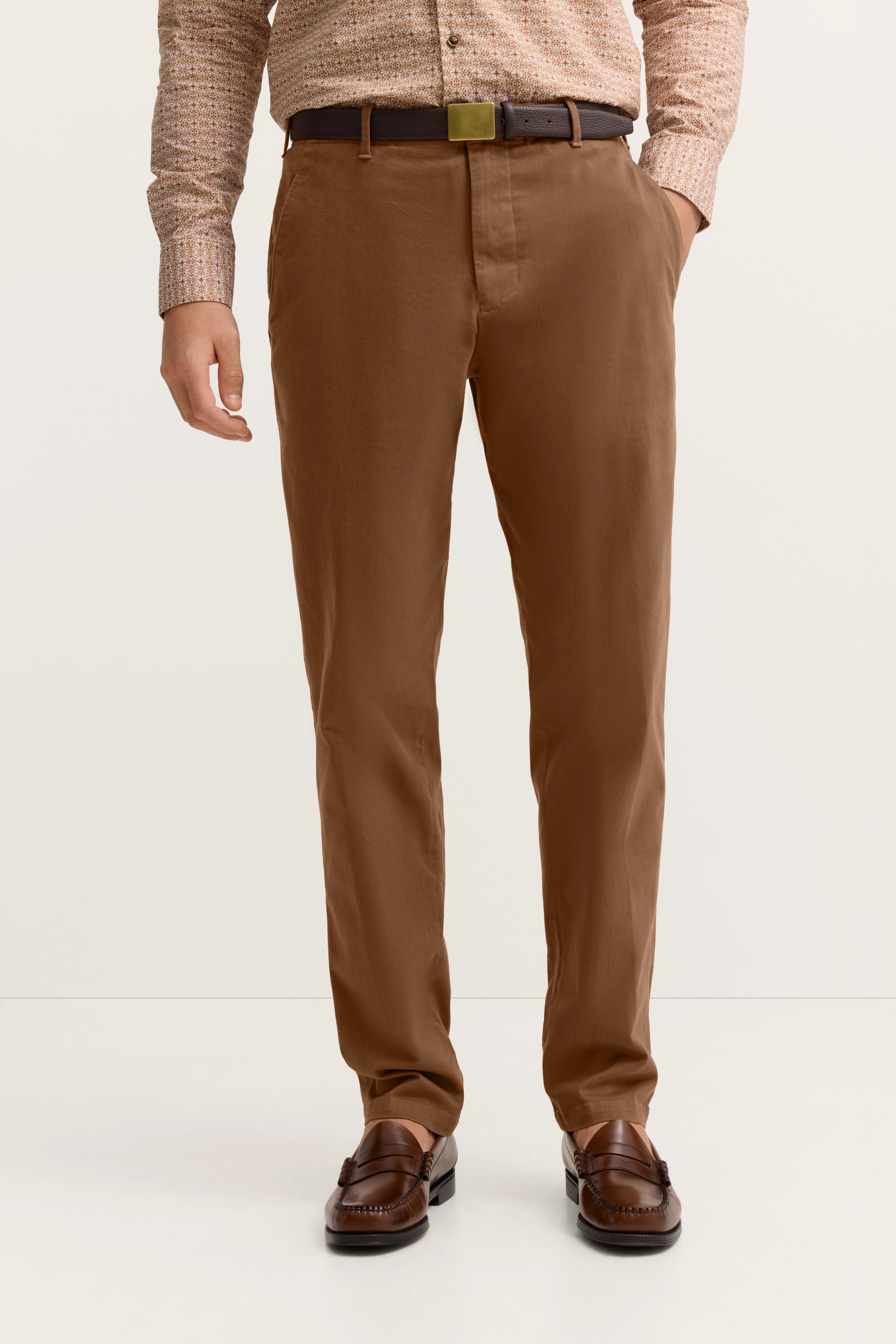 Braune Chinohose, gemustertes Hemd, dunkler Gürtel, Loafer. Büstenbild.