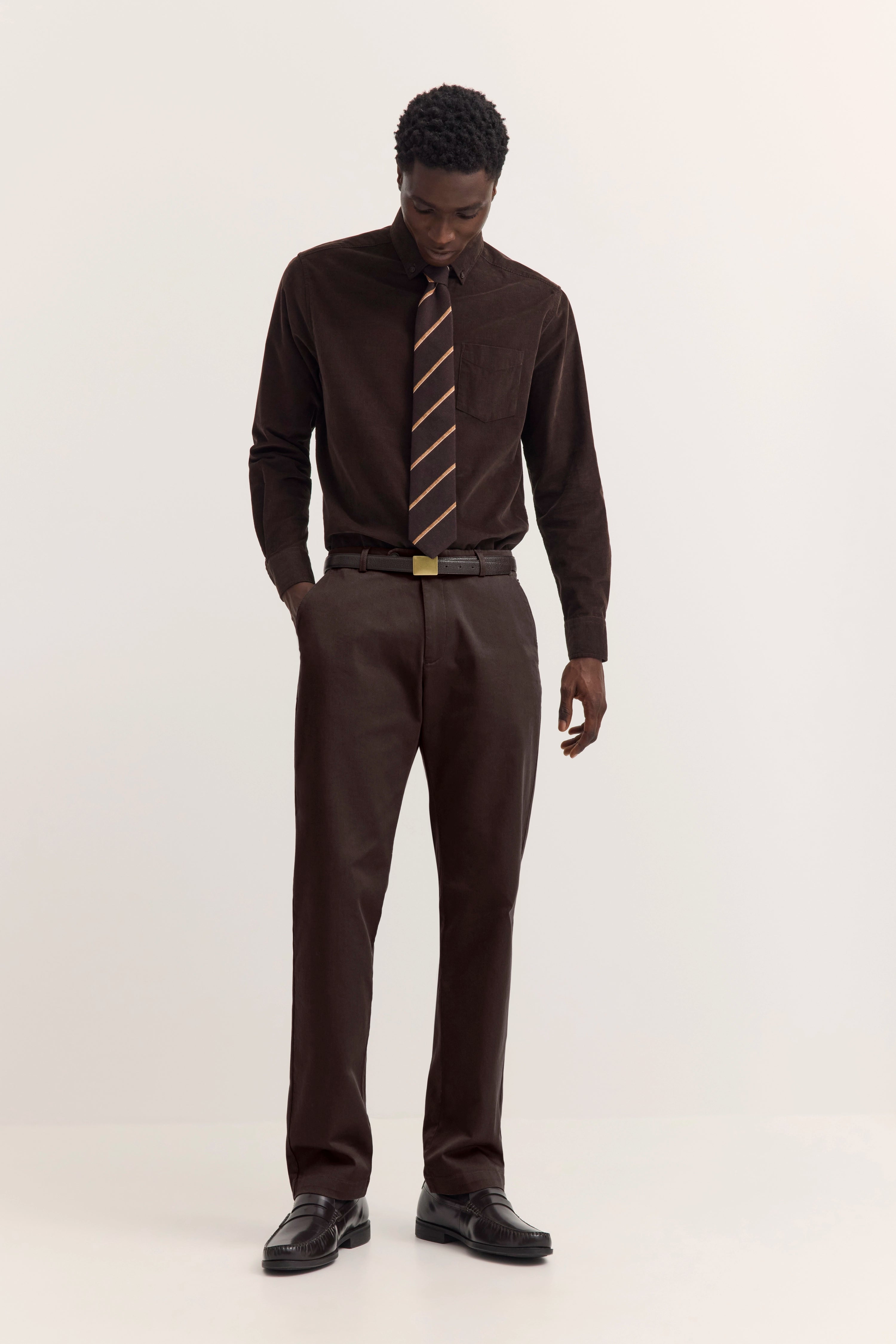 Model in braunem Hemd mit gestreifter Krawatte, brauner Hose und schwarzen Schuhen, entspanntes Posing.