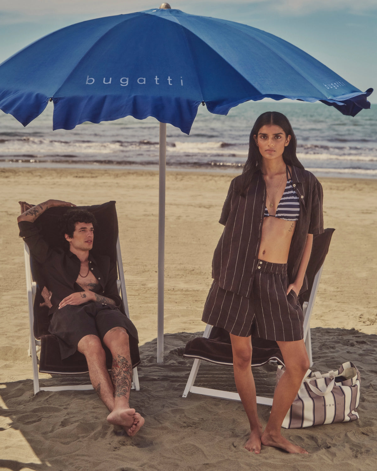 bugatti Strandoutfit: gestreifter Bikini, braunes Hemd und Shorts. Model am Strand unter Schirm.