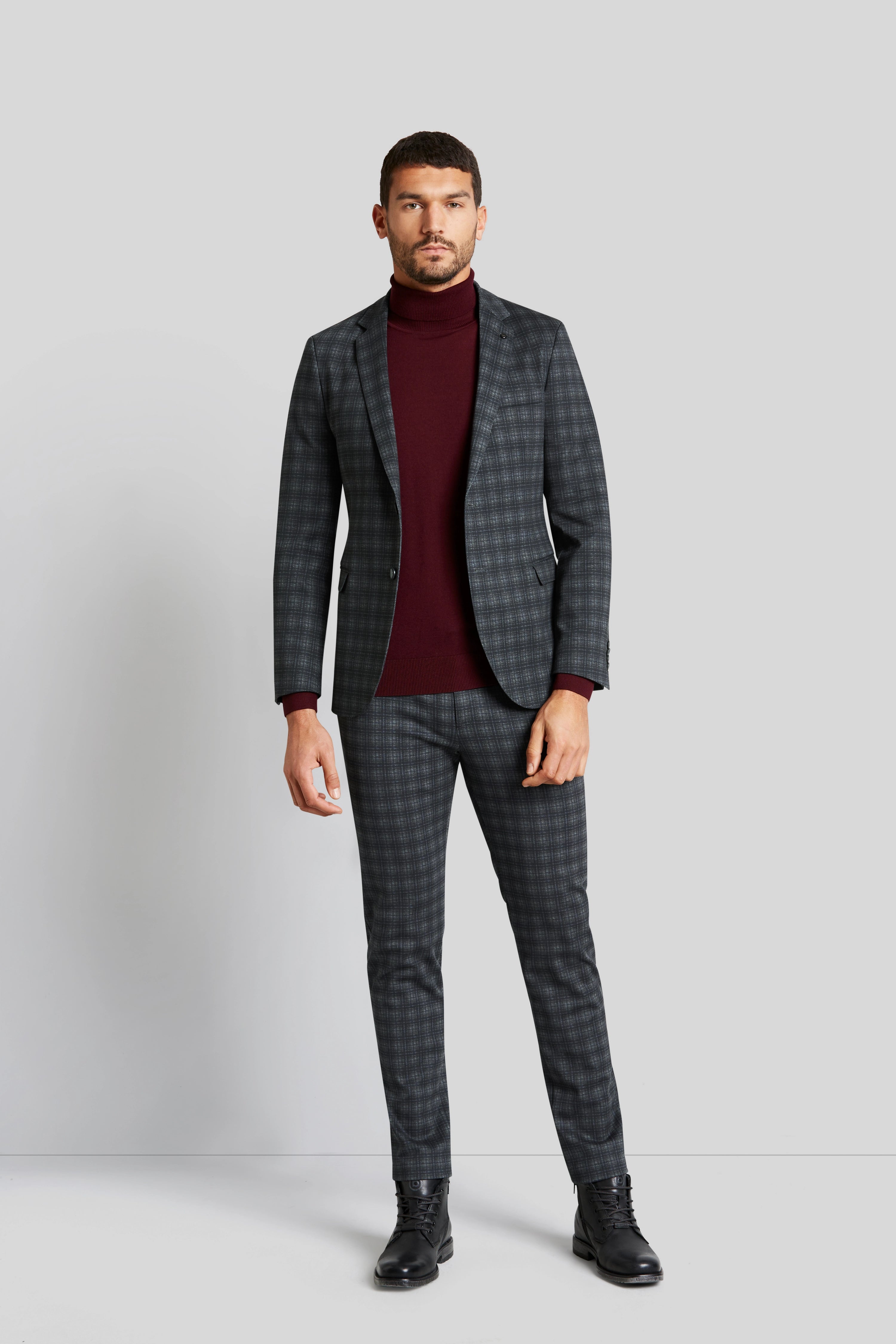 Man in checked suit with burgundy turtleneck, standing upright.