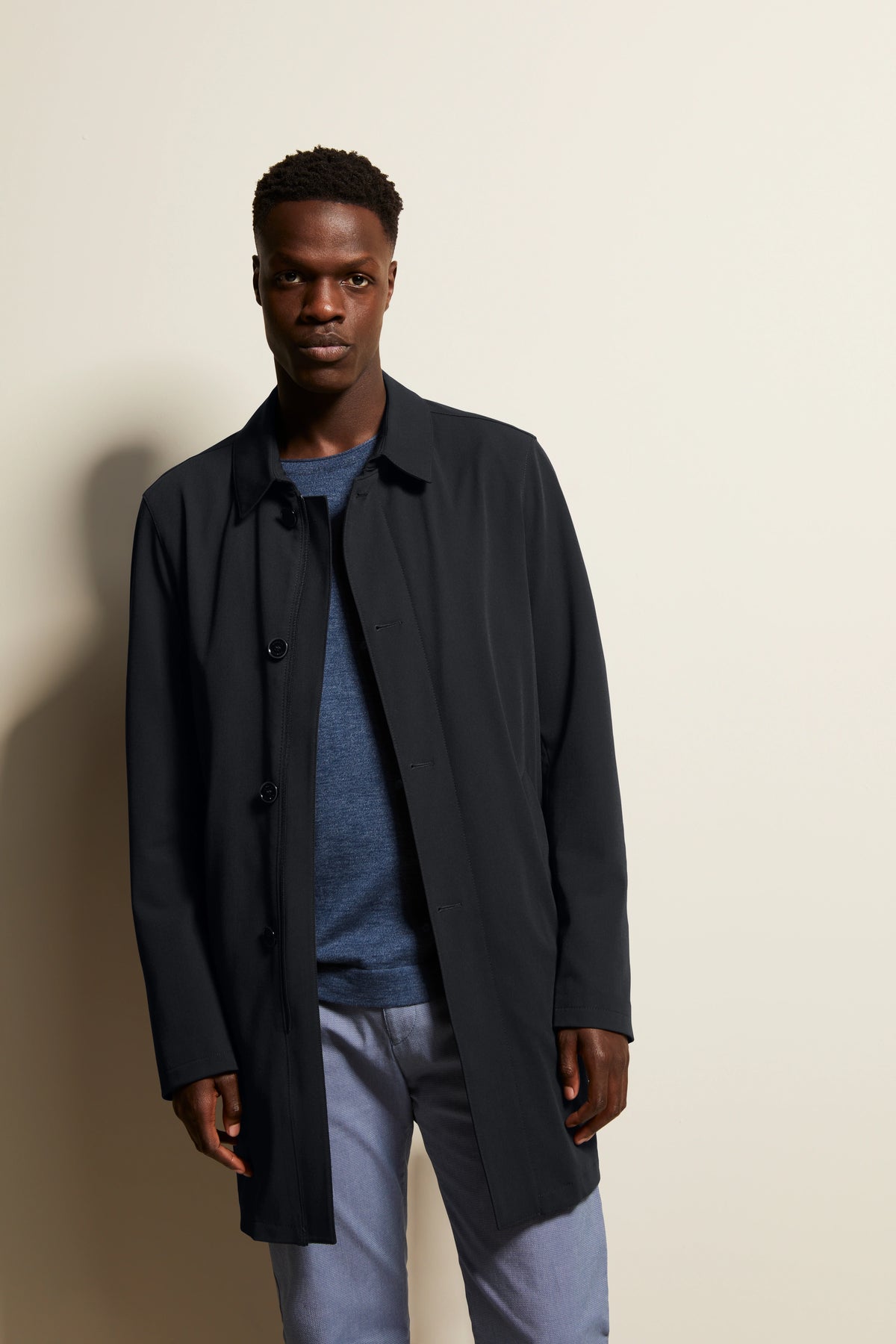 Model in black bugatti coat, blue shirt, gray pants, standing relaxed.