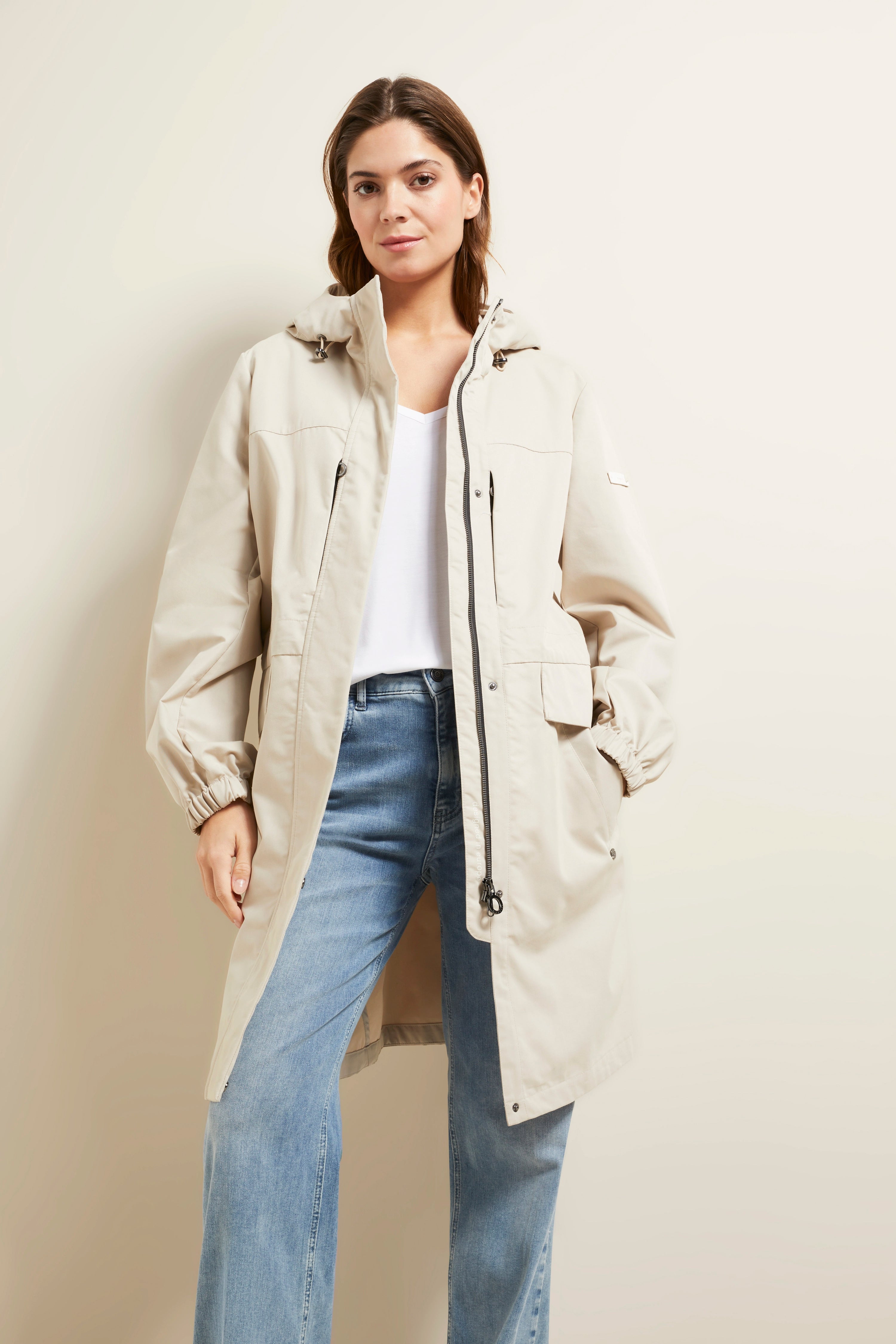 Beige bugatti coat with hood, white top, blue jeans, model standing, casually relaxed pose.