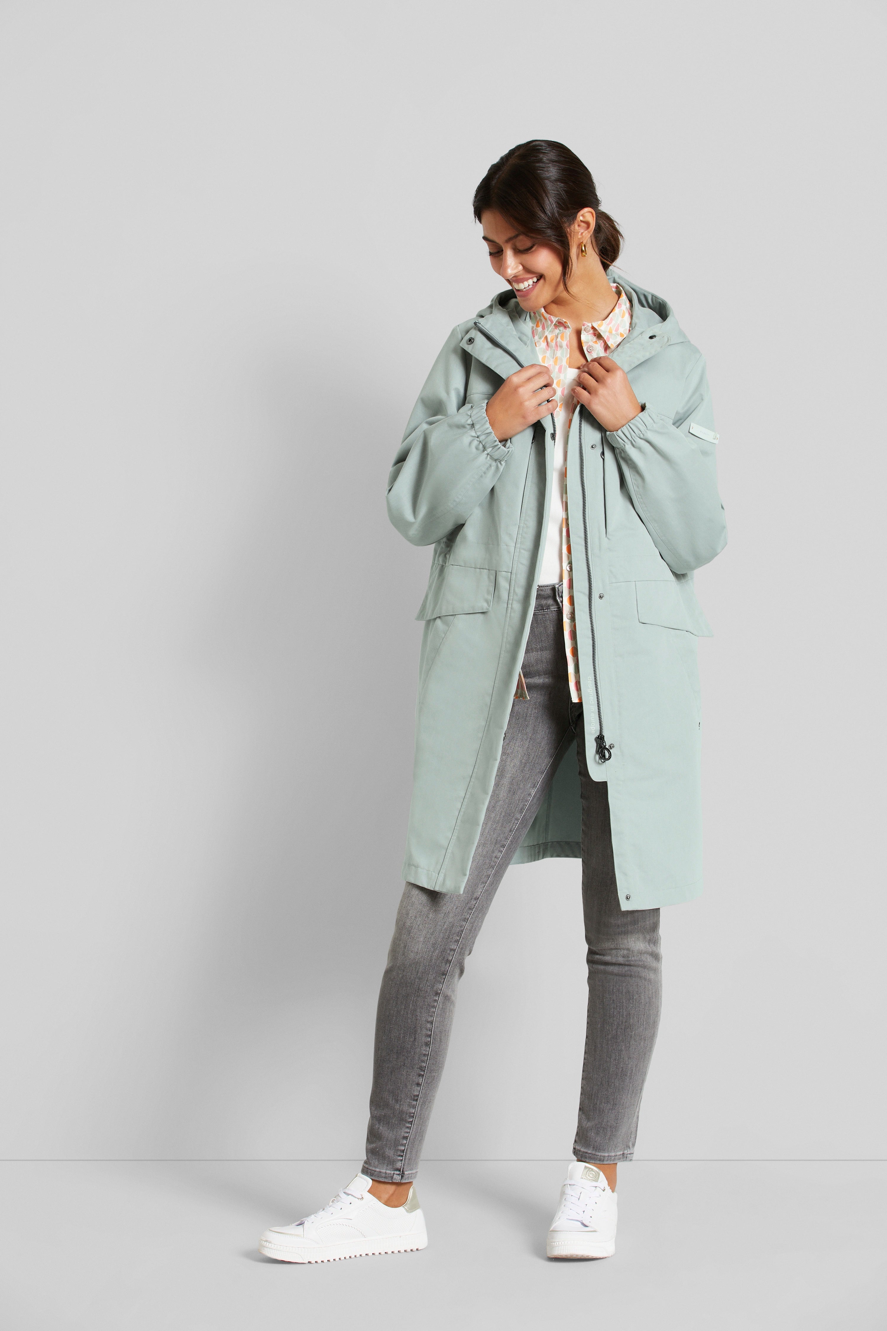 Model in hellblauer bugatti-Jacke, gemusterter Bluse und grauer Jeans, lässig stehend mit lächelndem Blick.